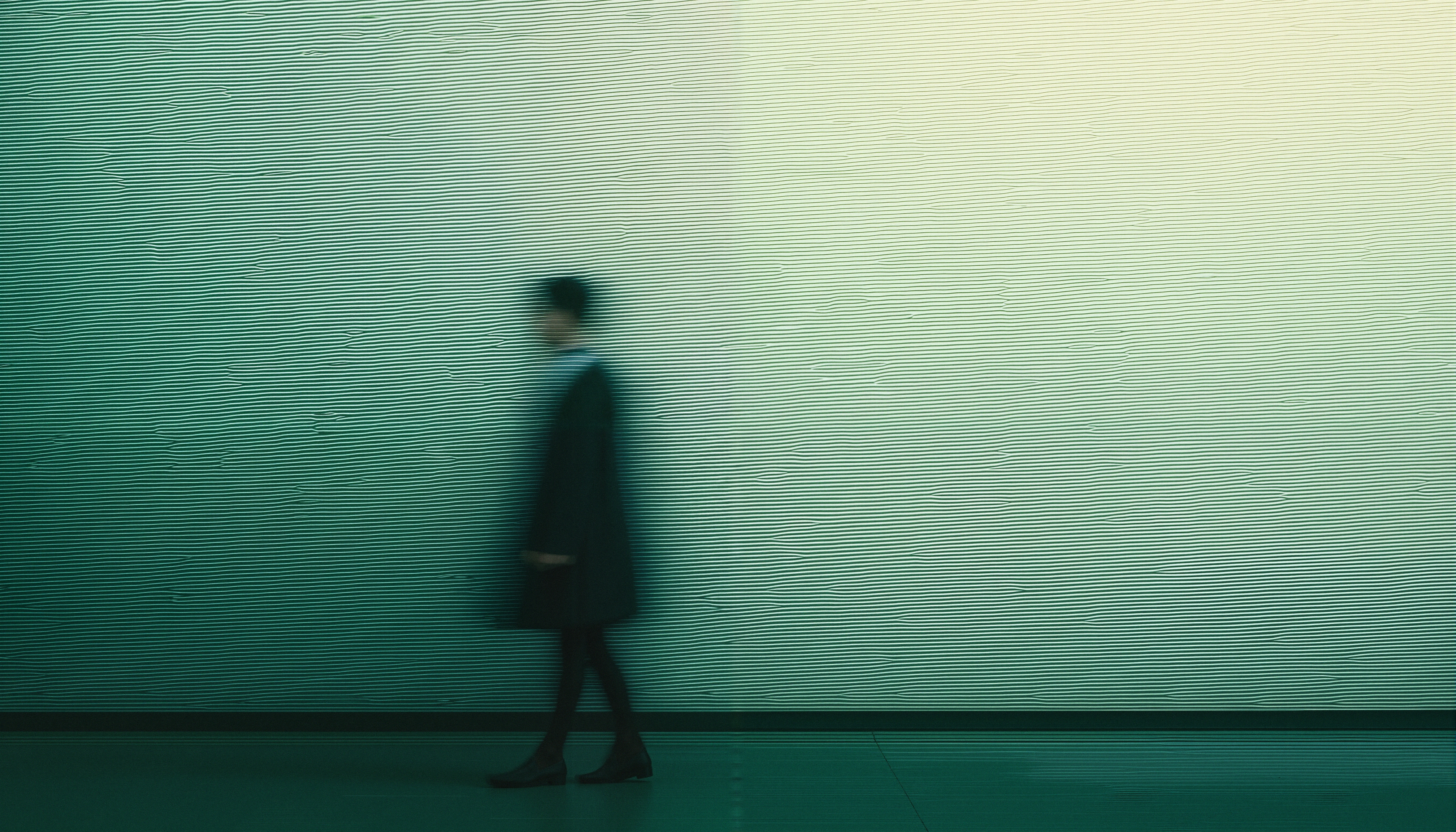 Blurred figure walking past a textured, greenish wall with linear details.