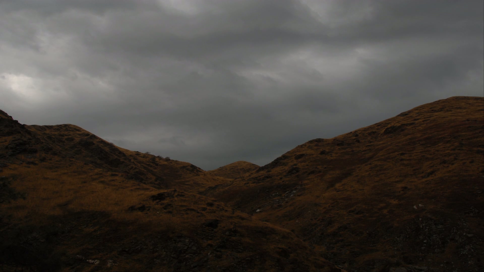 An atmospheric video of rugged, brown hills under a time-lapse, overcast sky, showcasing the stark beauty and desolate charm of the mountainous landscape.