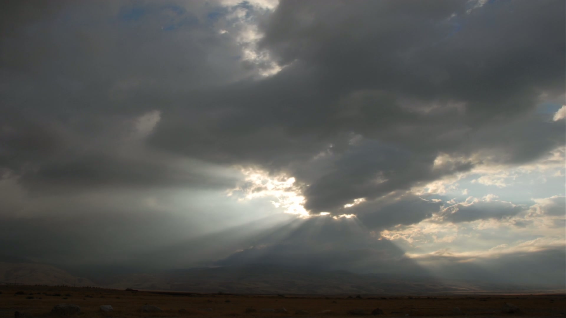 A dramatic time-lapse sky filled with dark clouds, highlighted by sunbeams breaking through, casts light over an arid plain with scattered rocks, set against distant hills.