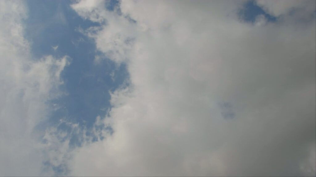 A scenic view of a time-lapse sky featuring a dramatic contrast between a clear blue area with wispy white clouds and dense greyish-white clouds, suggesting changing weather conditions.