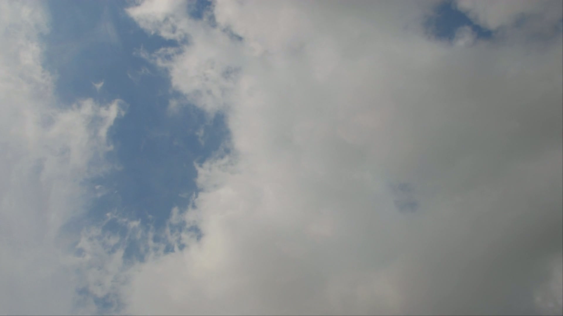 A scenic view of a time-lapse sky featuring a dramatic contrast between a clear blue area with wispy white clouds and dense greyish-white clouds, suggesting changing weather conditions.