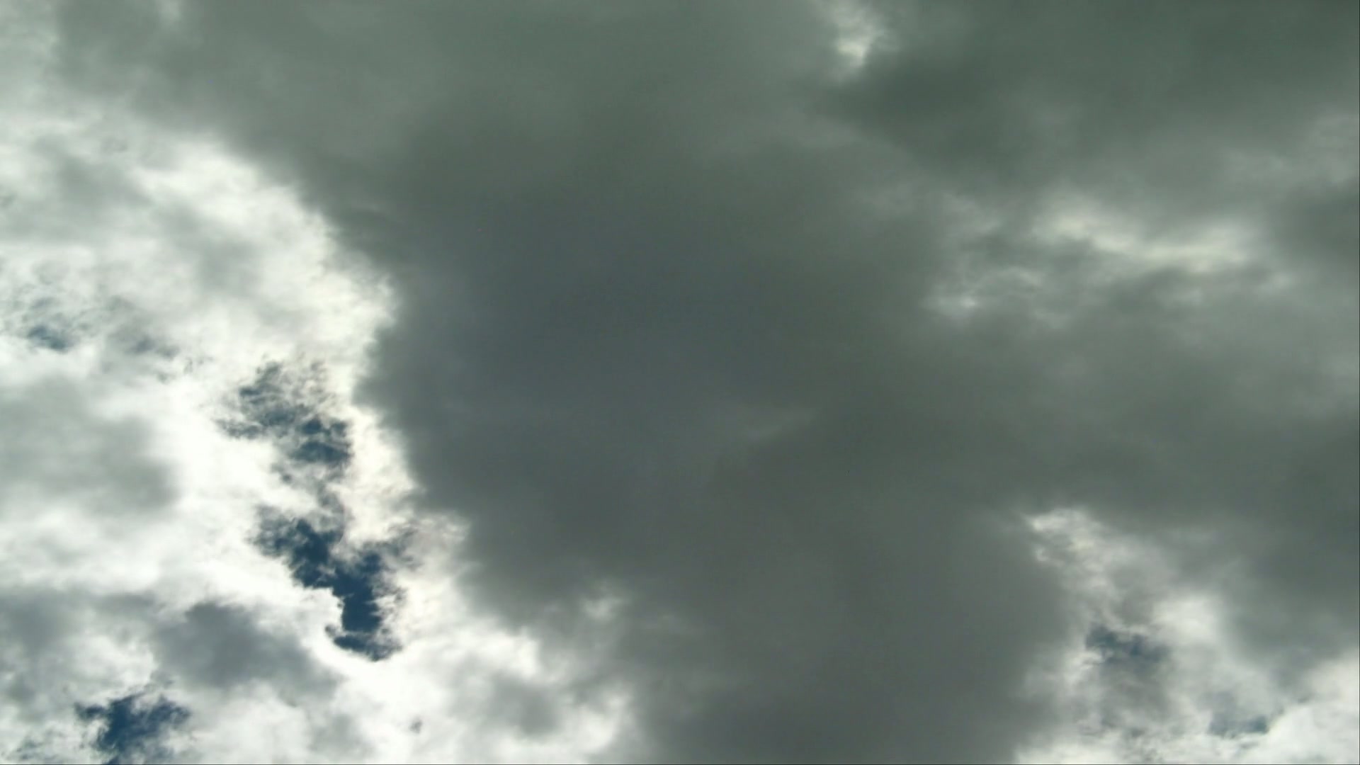Dramatic time-lapse cloudy sky with dark, ominous clouds overshadowing the scene, contrasting with lighter patches and glimpses of blue sky, suggesting an upcoming storm.