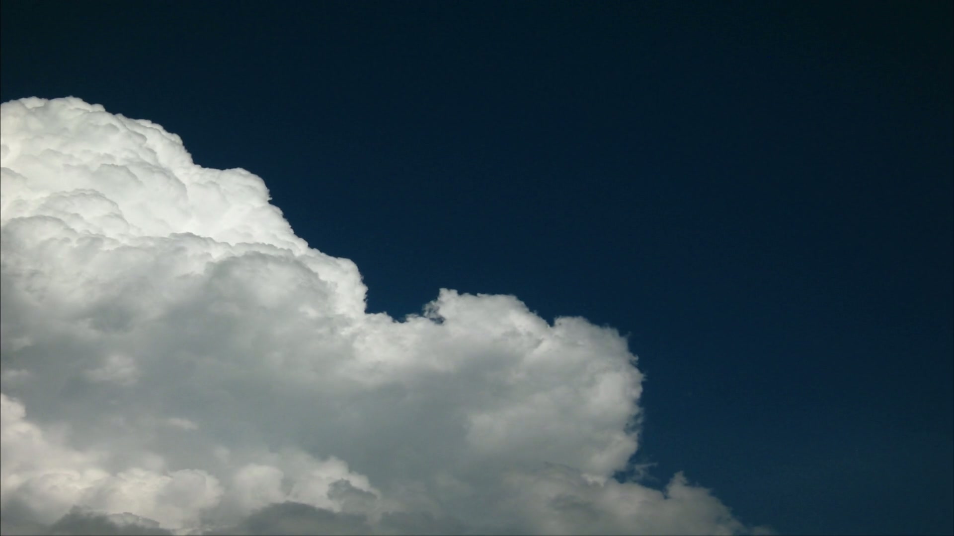 A large, white fluffy time-lapse cloud set against a deep blue sky, illuminated by sunlight, creating a peaceful and serene natural scene.