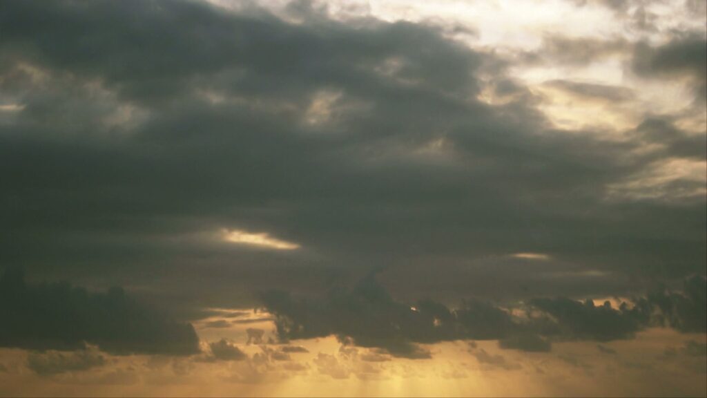 Dramatic sky filled with dark time-lapse clouds and beams of sunlight peeking through, casting a golden hue over the horizon, creating a striking contrast and serene yet foreboding atmosphere.