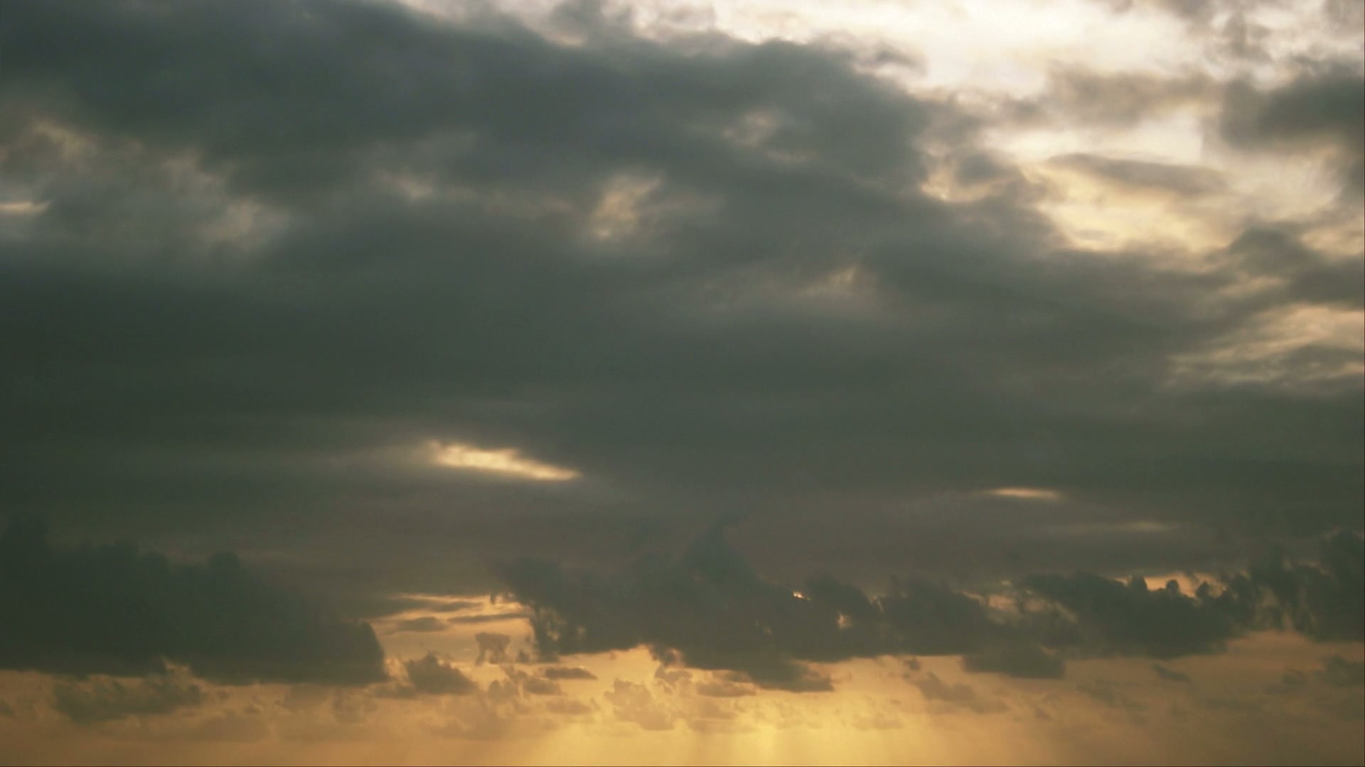 Dramatic sky filled with dark time-lapse clouds and beams of sunlight peeking through, casting a golden hue over the horizon, creating a striking contrast and serene yet foreboding atmosphere.
