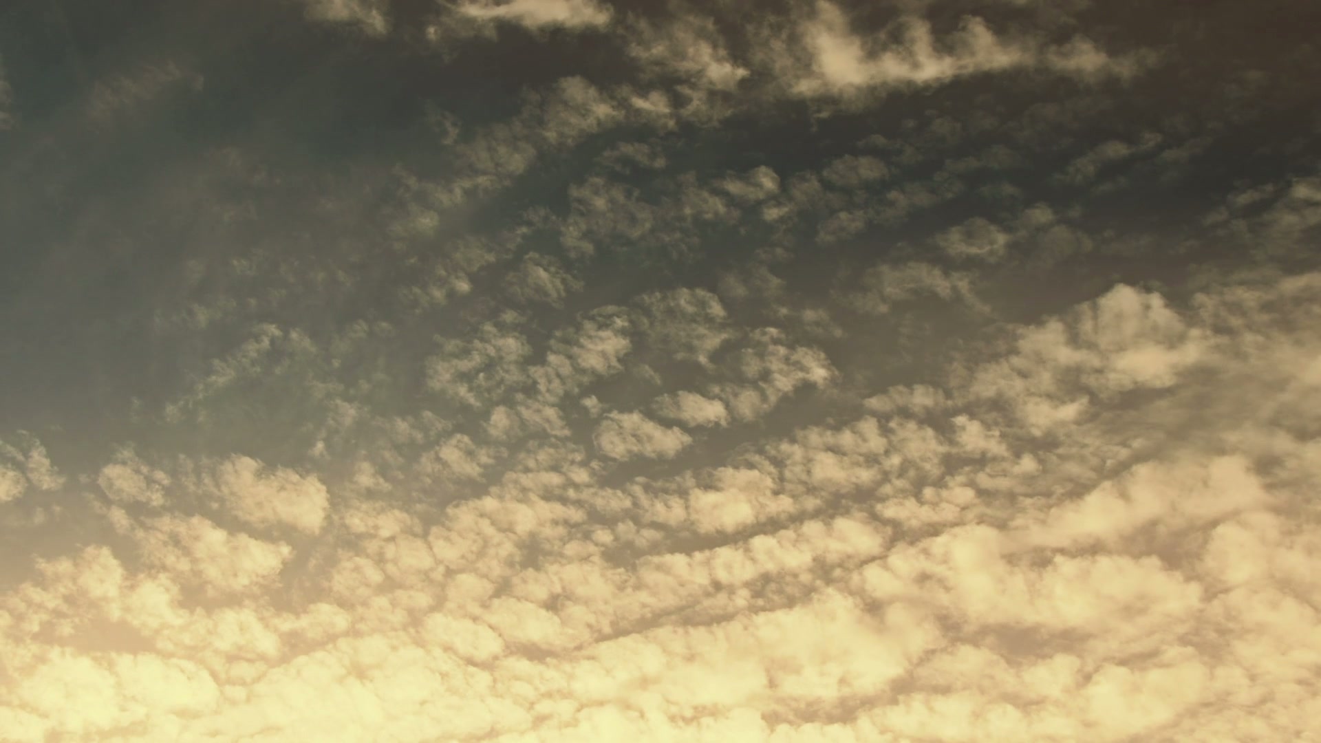 A peaceful sky filled with scattered, fluffy time-lapse clouds transitioning from a golden hue at the bottom to a darker shade above, creating a serene atmosphere during sunrise or sunset.