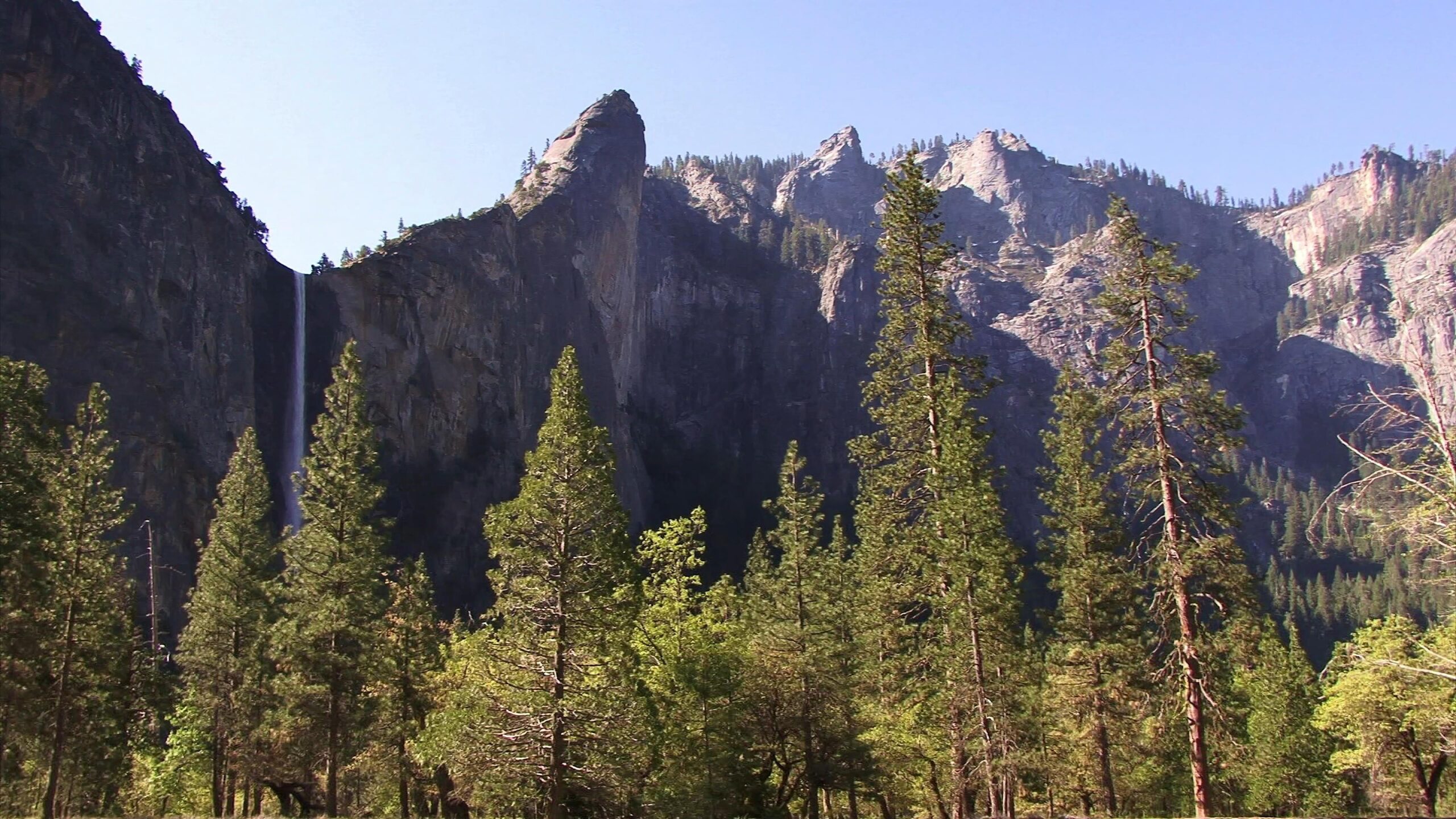Yosemite Falls surrounded by pine trees and towering granite cliffs on sunny day