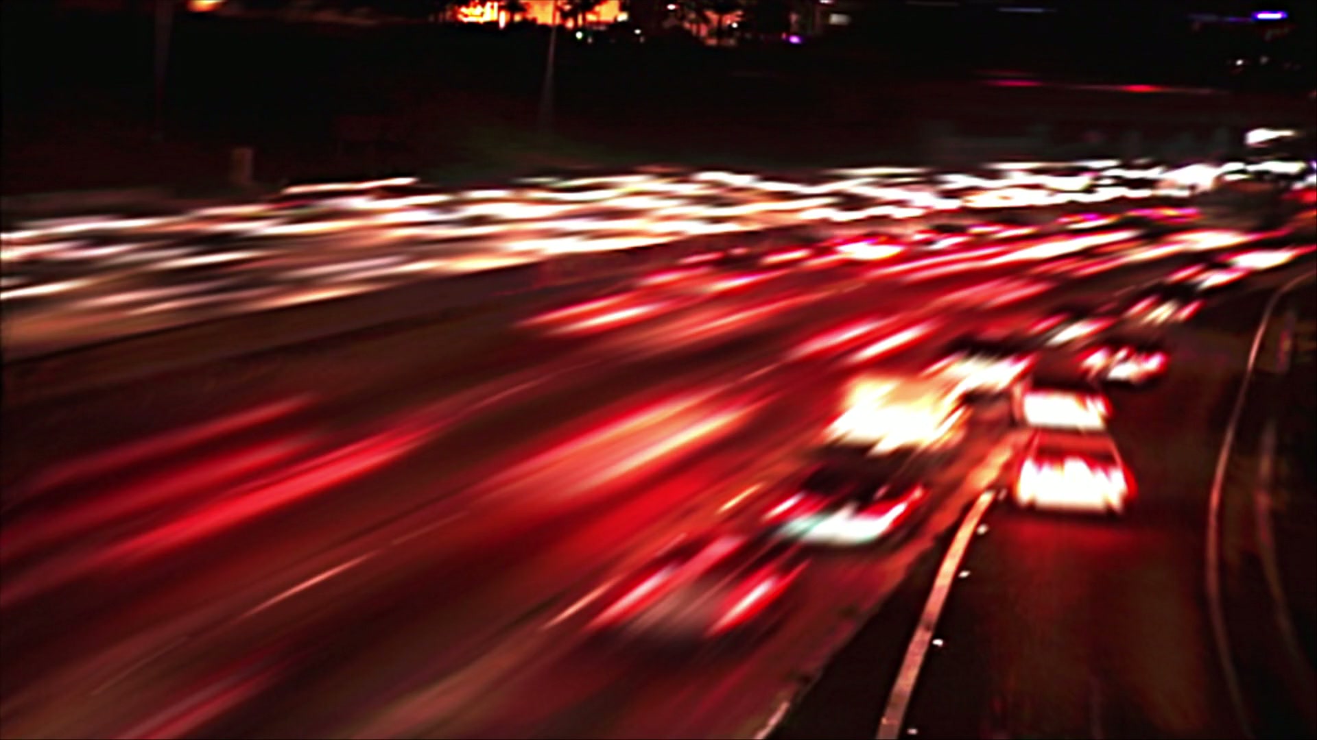 A looping video of a dynamic nighttime highway scene with long exposure captures colorful light trails from fast-moving cars, set against a backdrop of city lights.