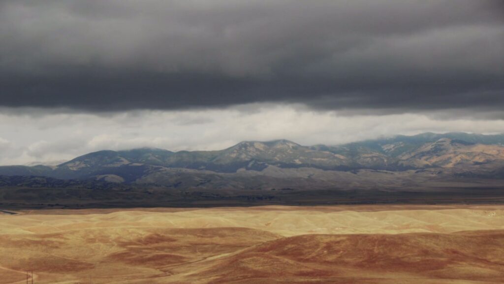 Dramatic landscape with golden rolling hills under stormy time-lapse clouds and distant mountains.