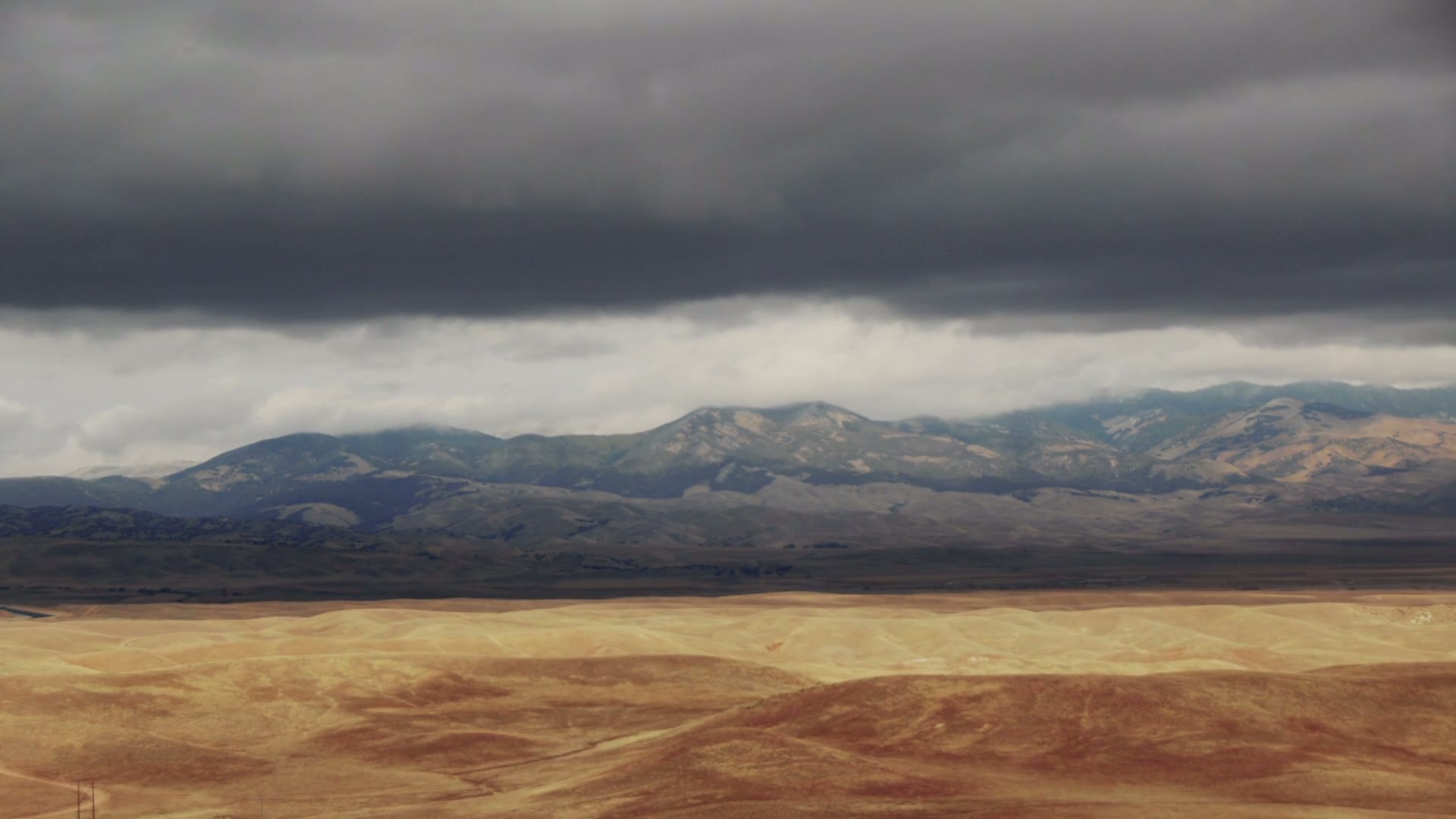 Dramatic landscape with golden rolling hills under stormy time-lapse clouds and distant mountains.