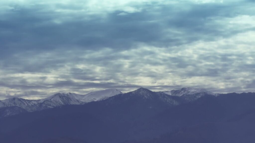 Serene mountain landscape with snow-capped peaks under a vast, time-lapse cloudy sky, evoking tranquility and natural beauty.