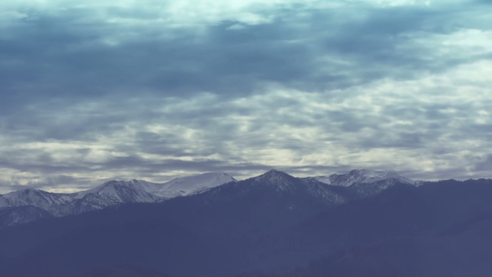 Serene mountain landscape with snow-capped peaks under a vast, time-lapse cloudy sky, evoking tranquility and natural beauty.