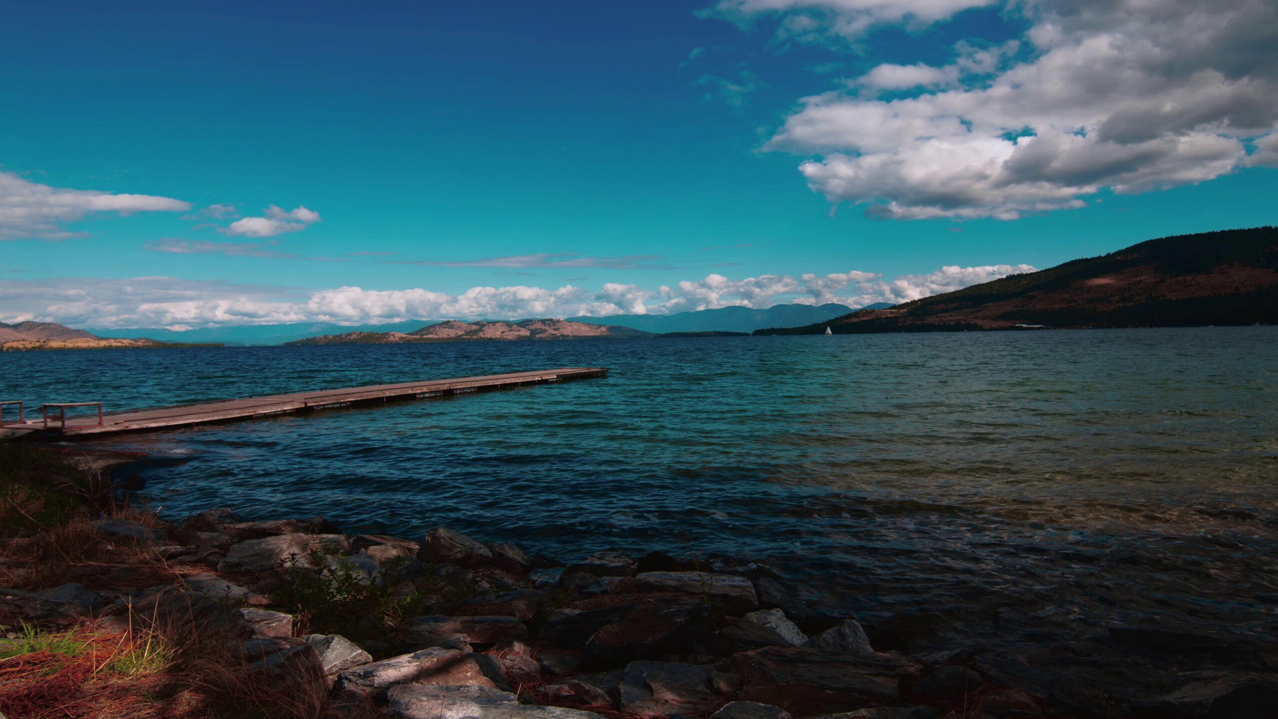 A serene lakeside scene with a wooden pier, surrounded by calm blue waters, distant hills, and a vibrant sky filled with fluffy time-lapse clouds.