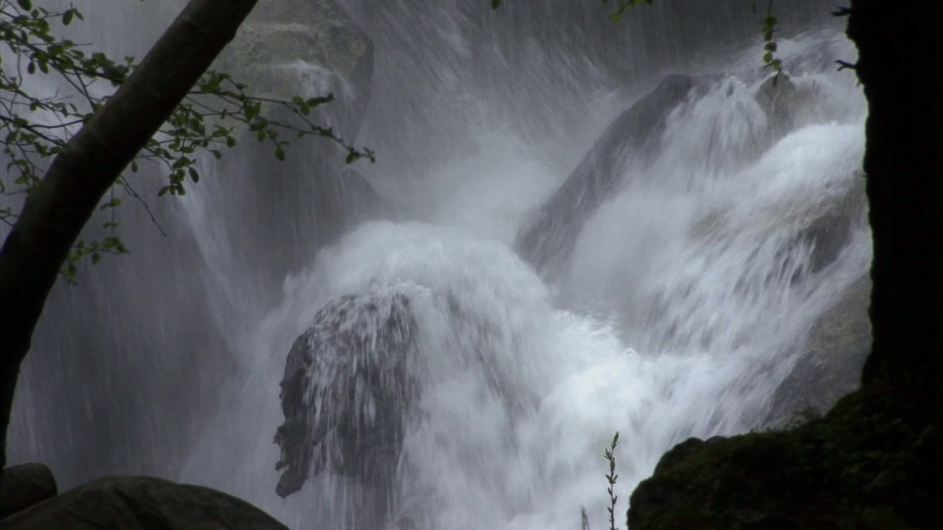 The video captures a powerful waterfall cascading over large rocks. Water is forcefully crashing down, creating a misty aura around the area.