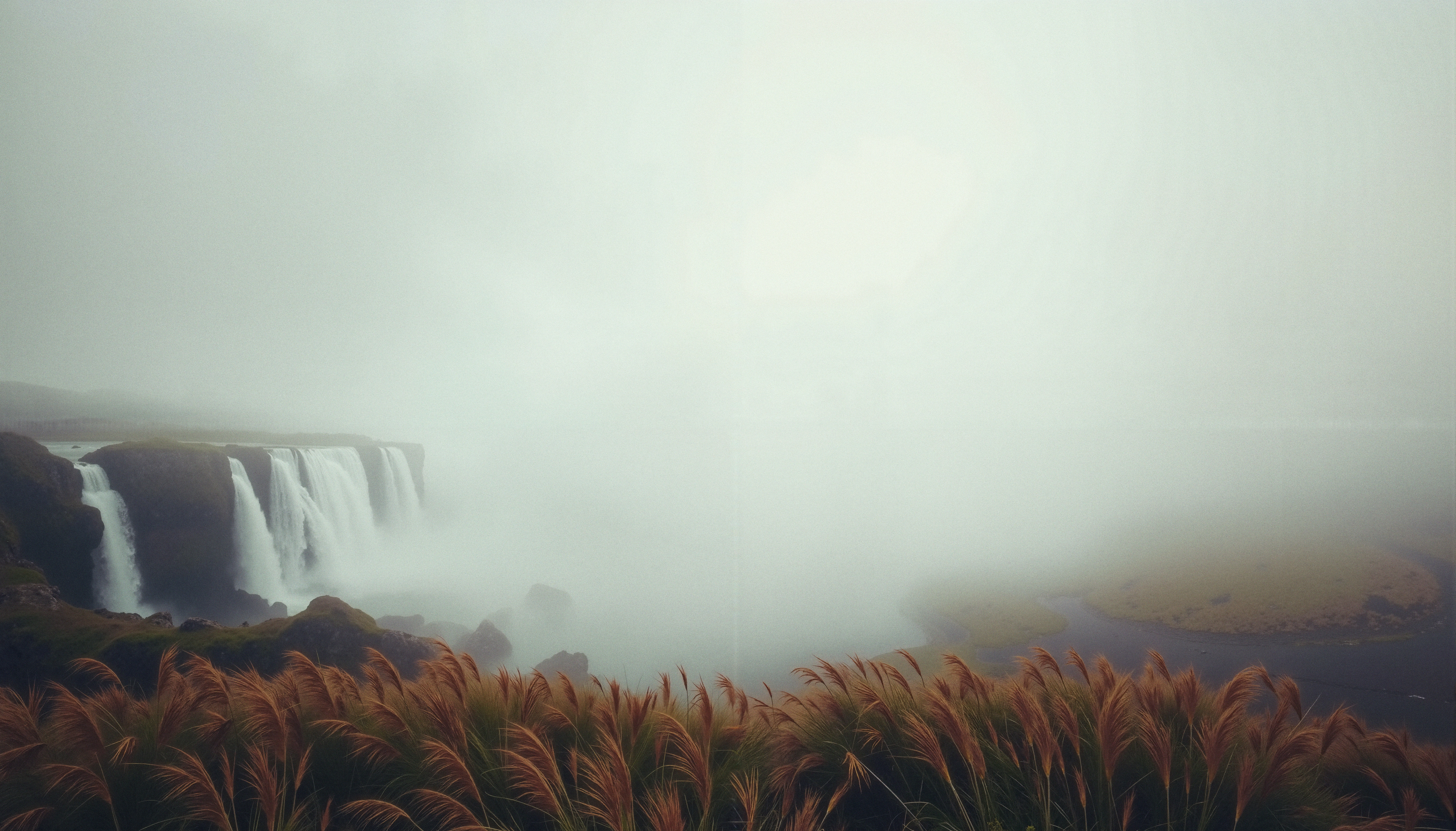 A misty waterfall surrounded by fog, cascading down rocky cliffs with swaying grasses in the foreground.
