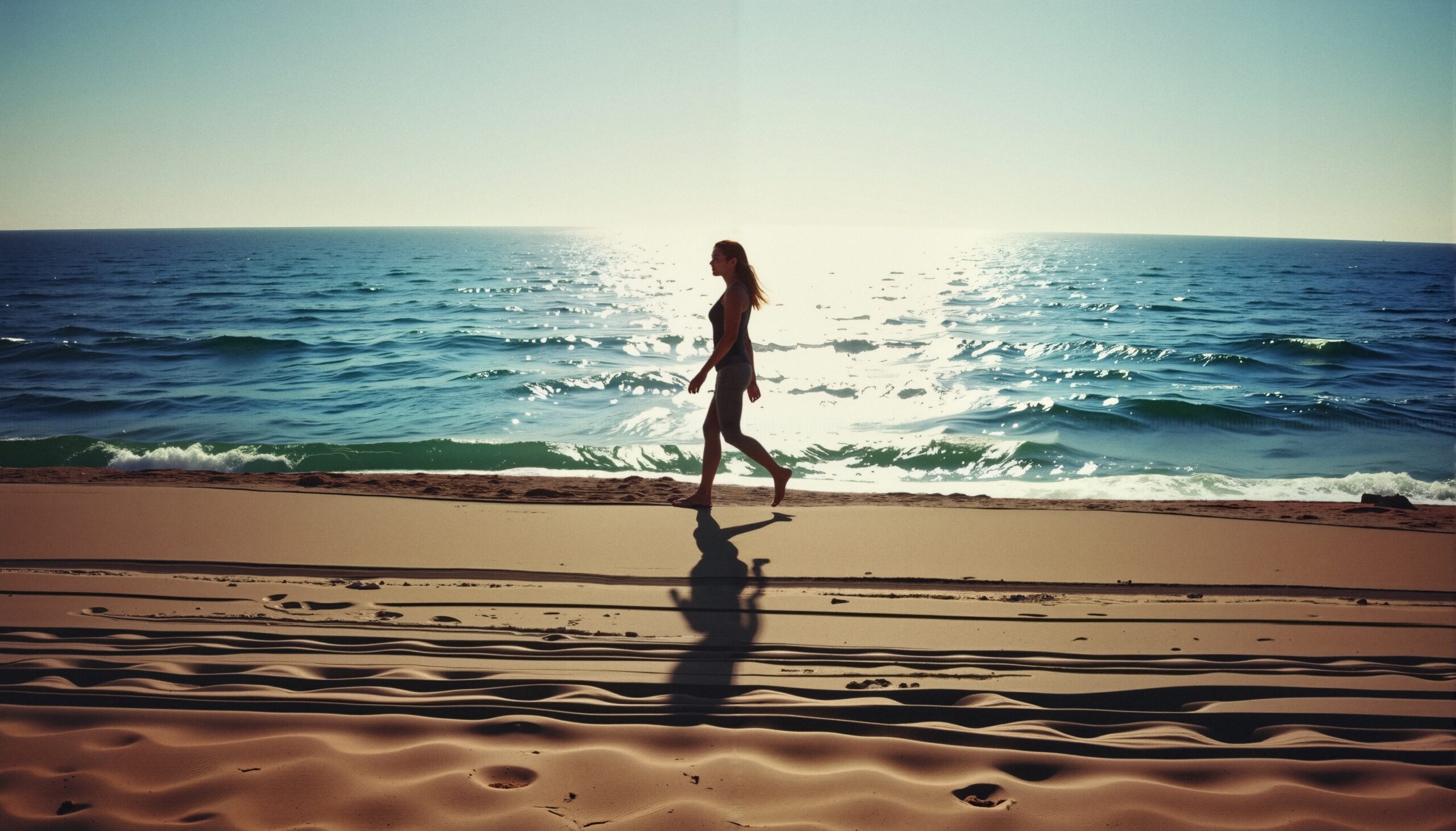 A woman walks along a sandy beach at sunset with shimmering ocean waves in the background.