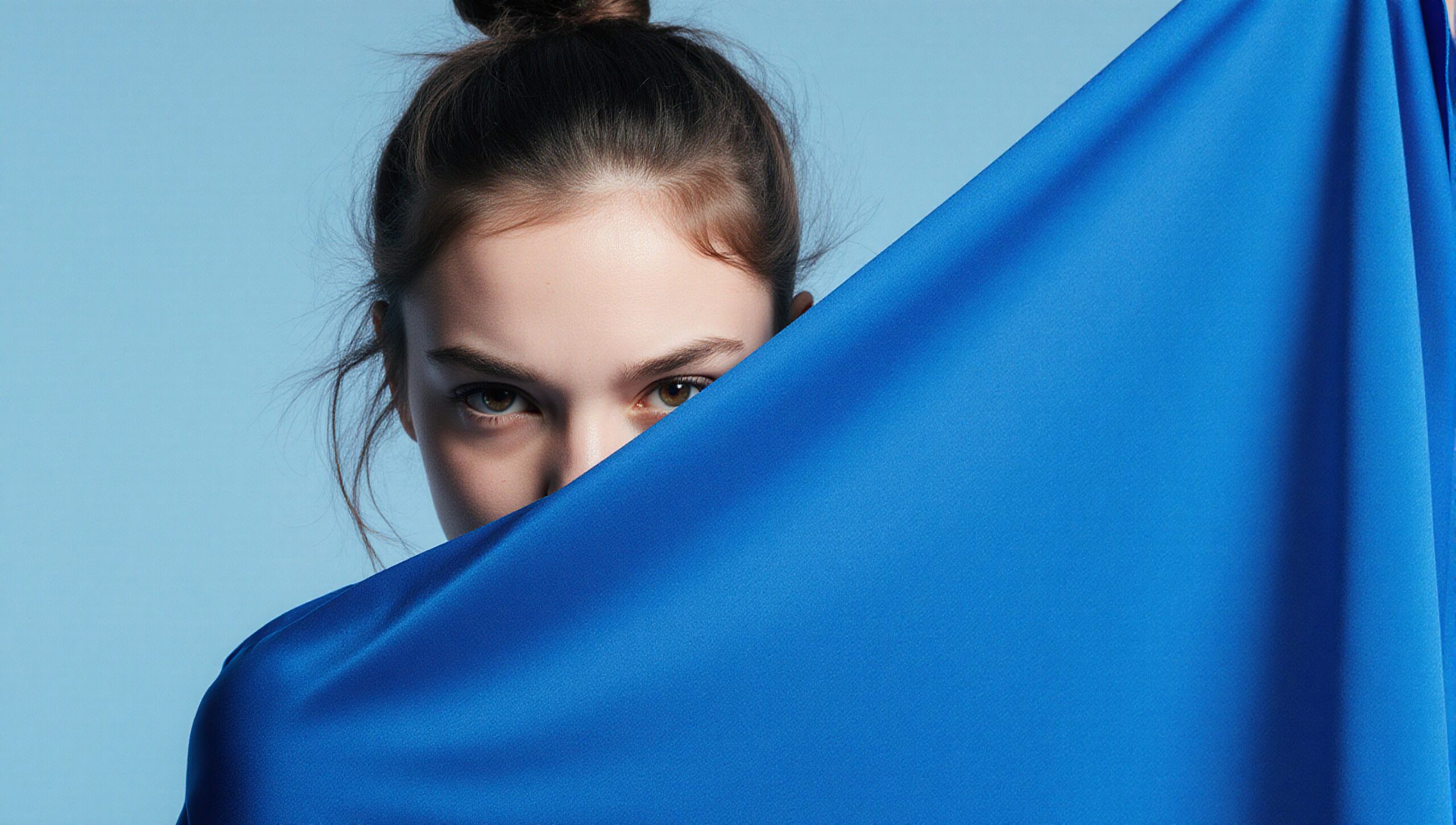 A woman with expressive eyes peers over a vibrant blue fabric in front of a light blue background.