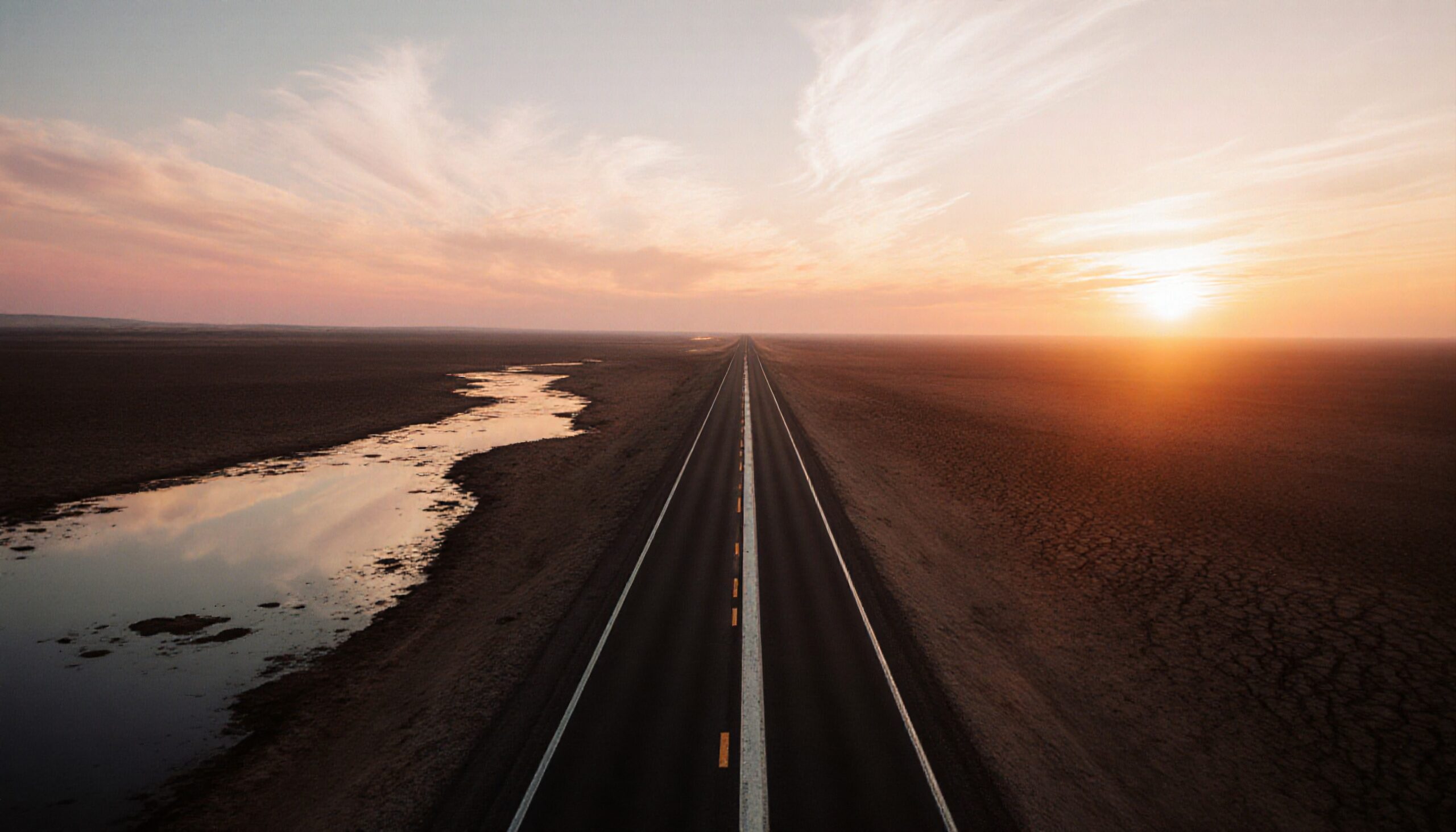 A long highway under a colorful sunset sky with reflections in the water and desert surroundings.