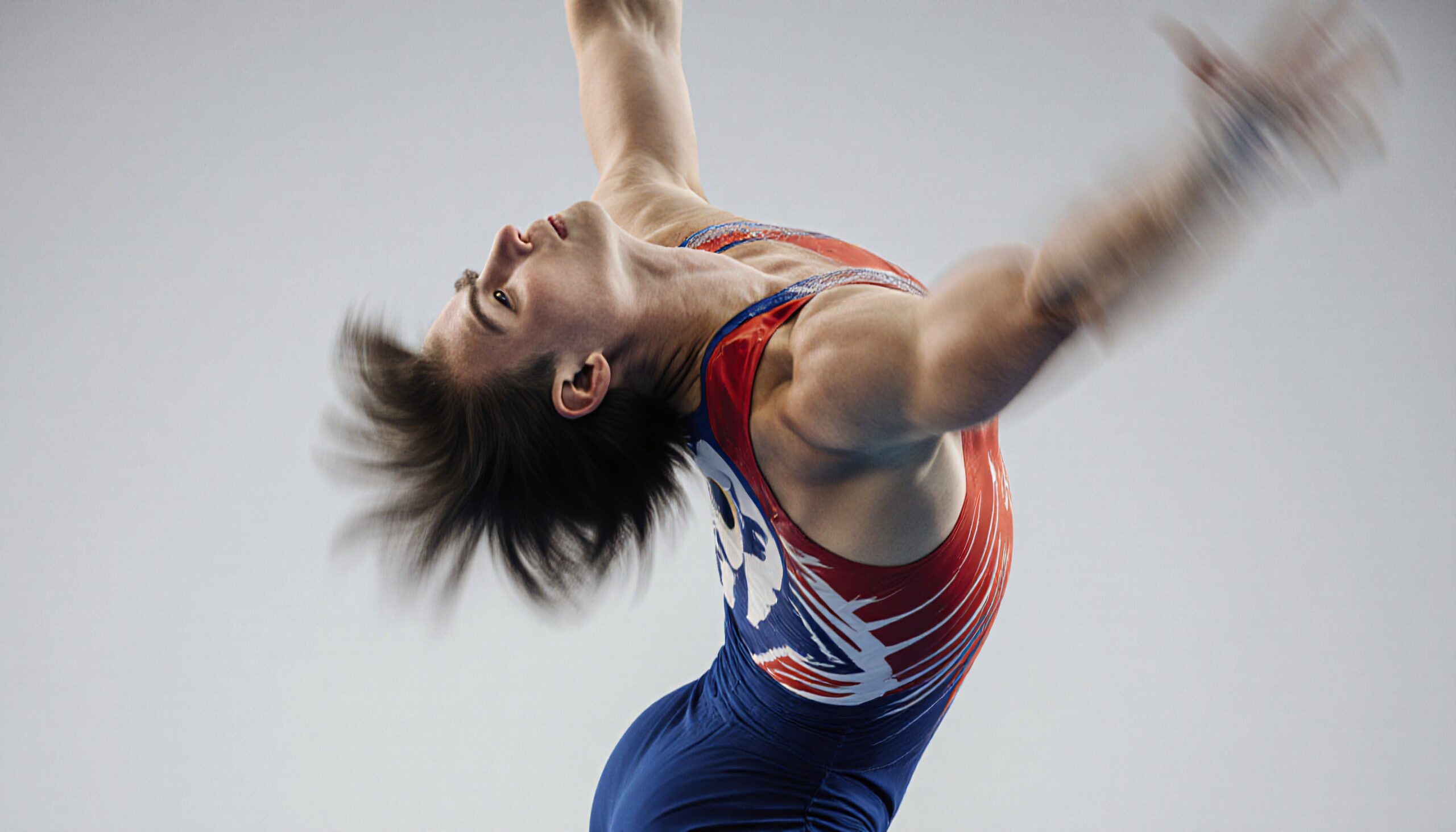 A gymnast in a colorful leotard performs a dynamic leap against a gray background.