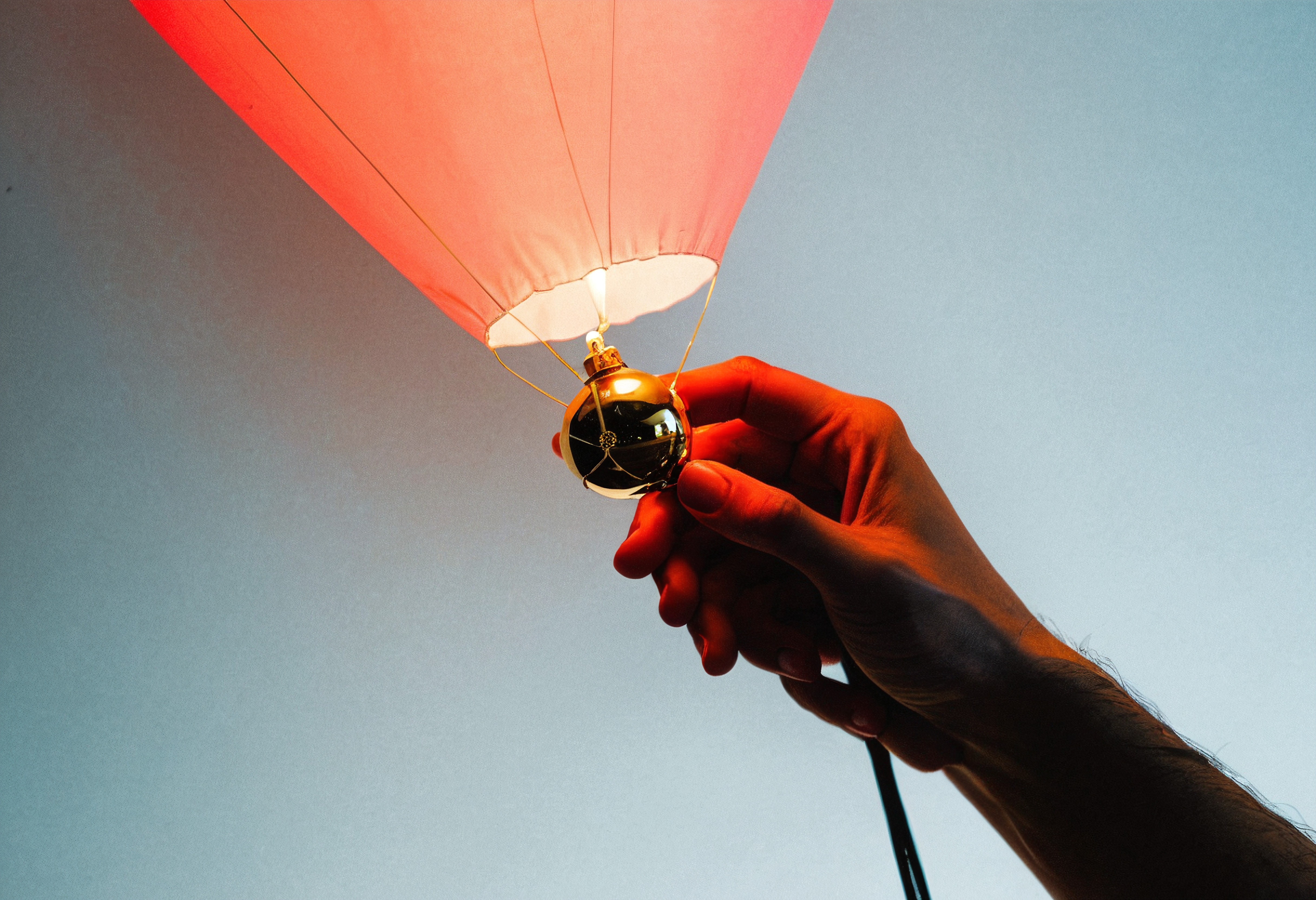 A hand holds a glowing red balloon base with blue sky.