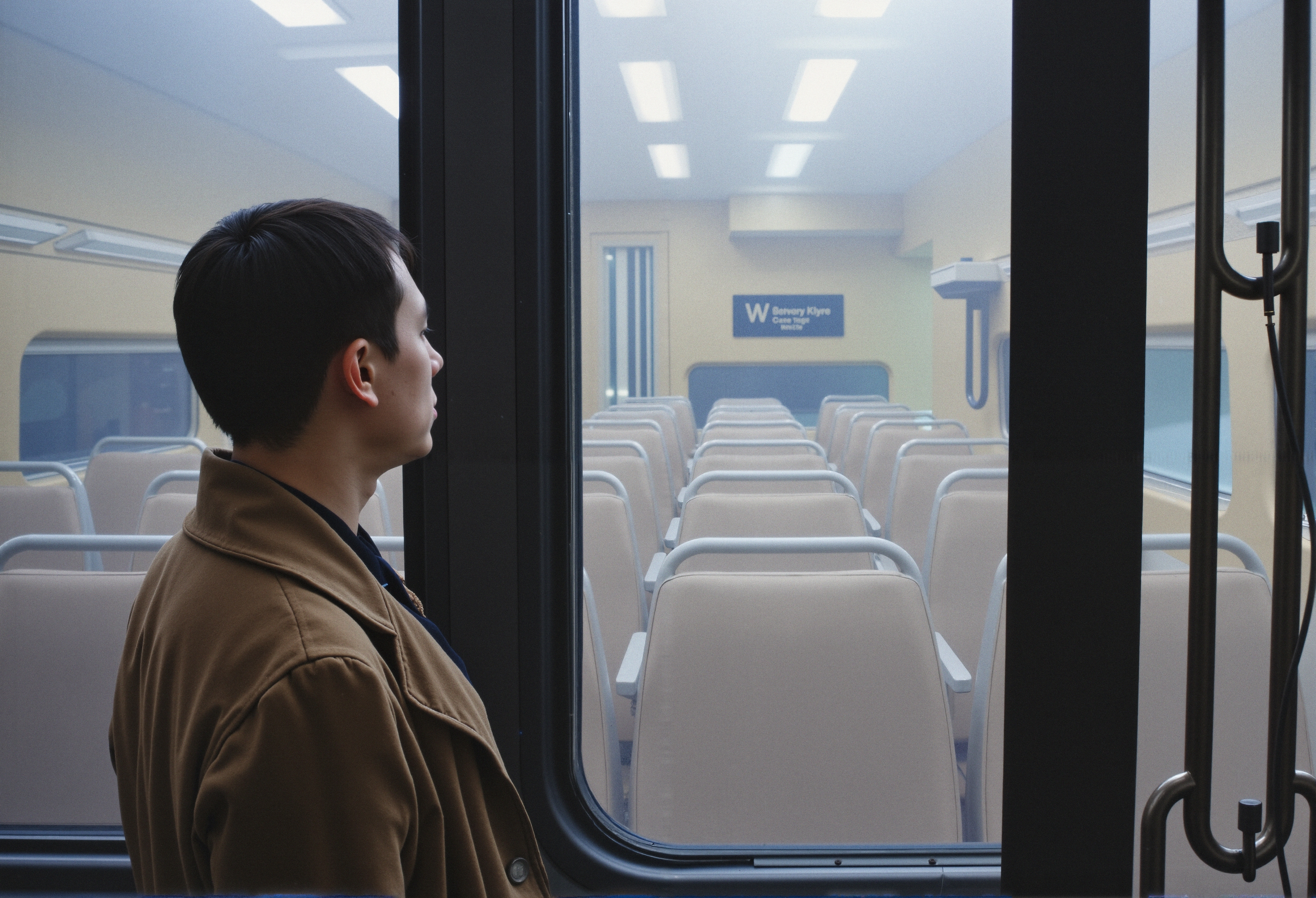 A person in a brown coat looks out of a train window, observing an empty adjacent car.