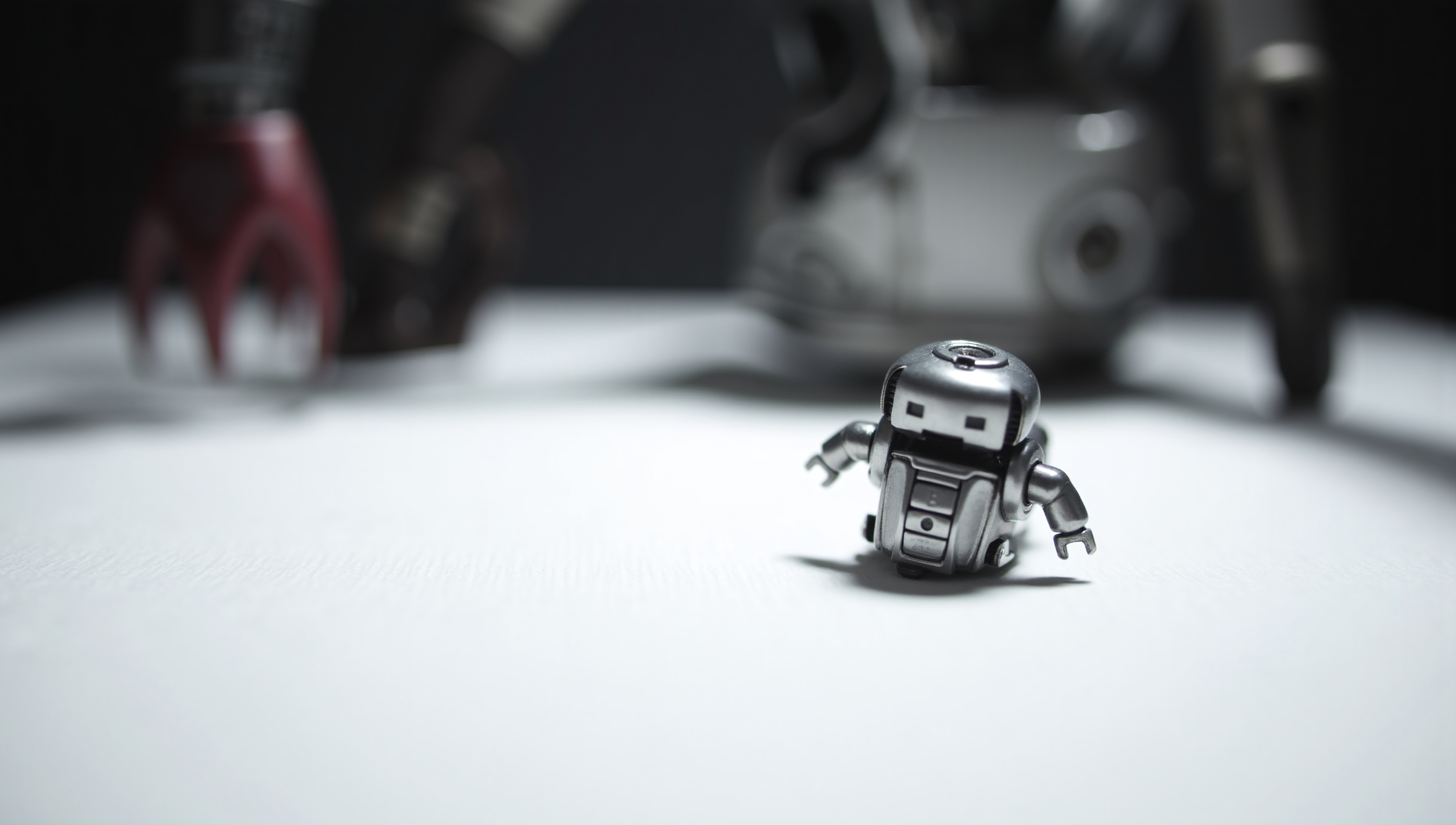 A small silver robot toy stands on a white surface surrounded by out-of-focus background elements.