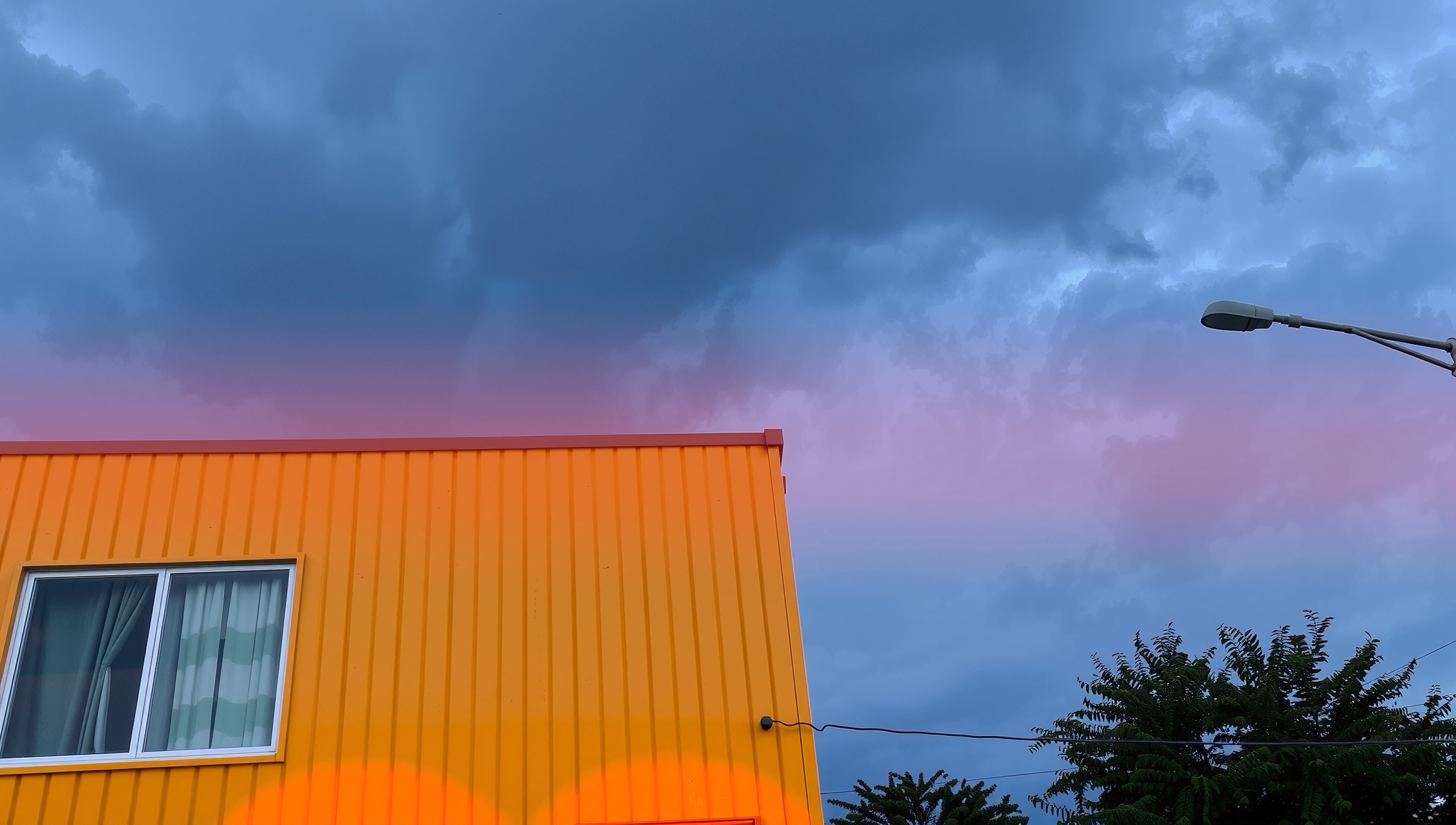 Vivid yellow building with corrugated metal exterior set against a dramatic sky, showcasing pink and purple cloud formations, with streetlamp and trees.