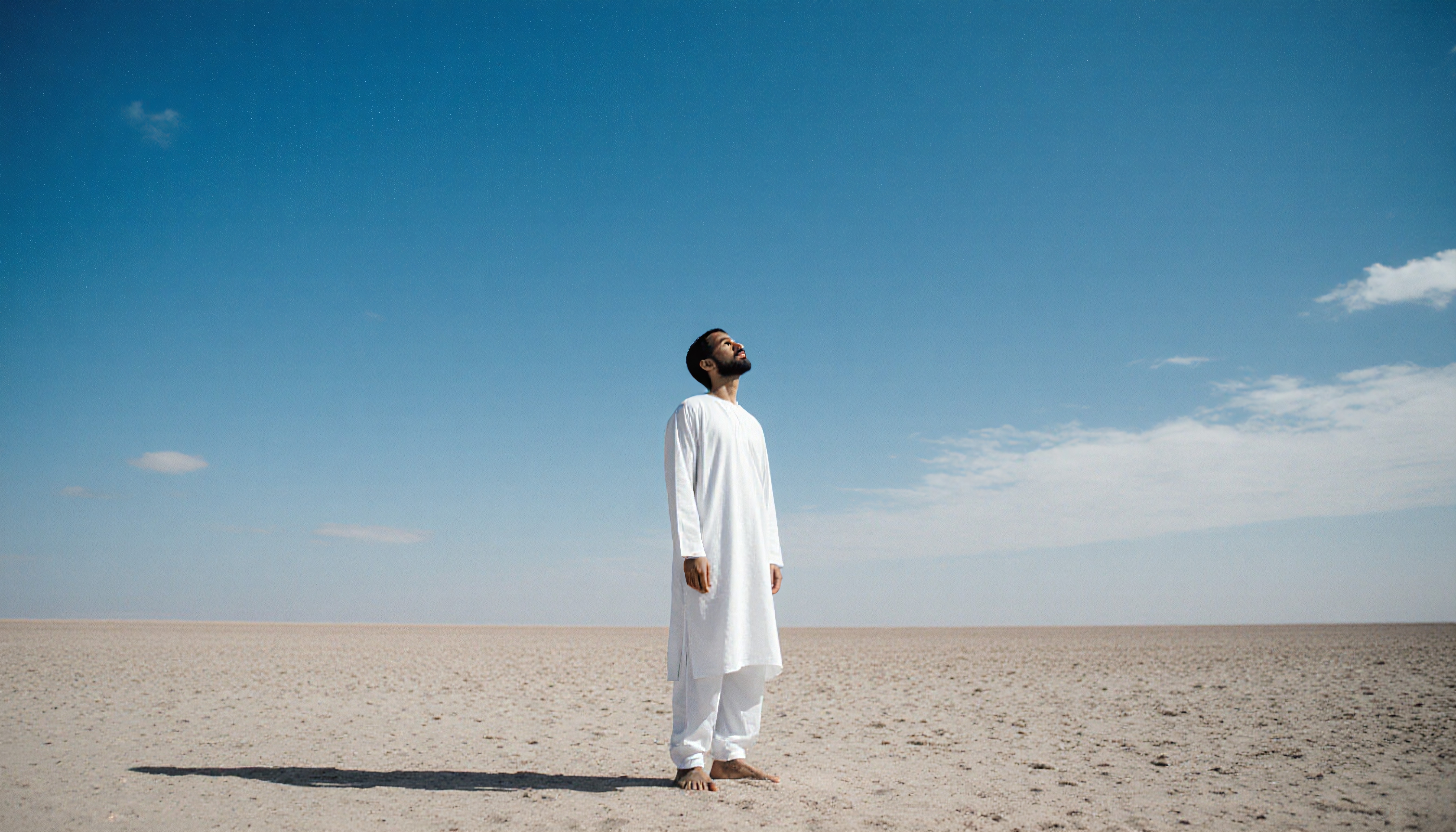 A man in white clothing stands barefoot in a vast desert, gazing thoughtfully at the open blue sky.