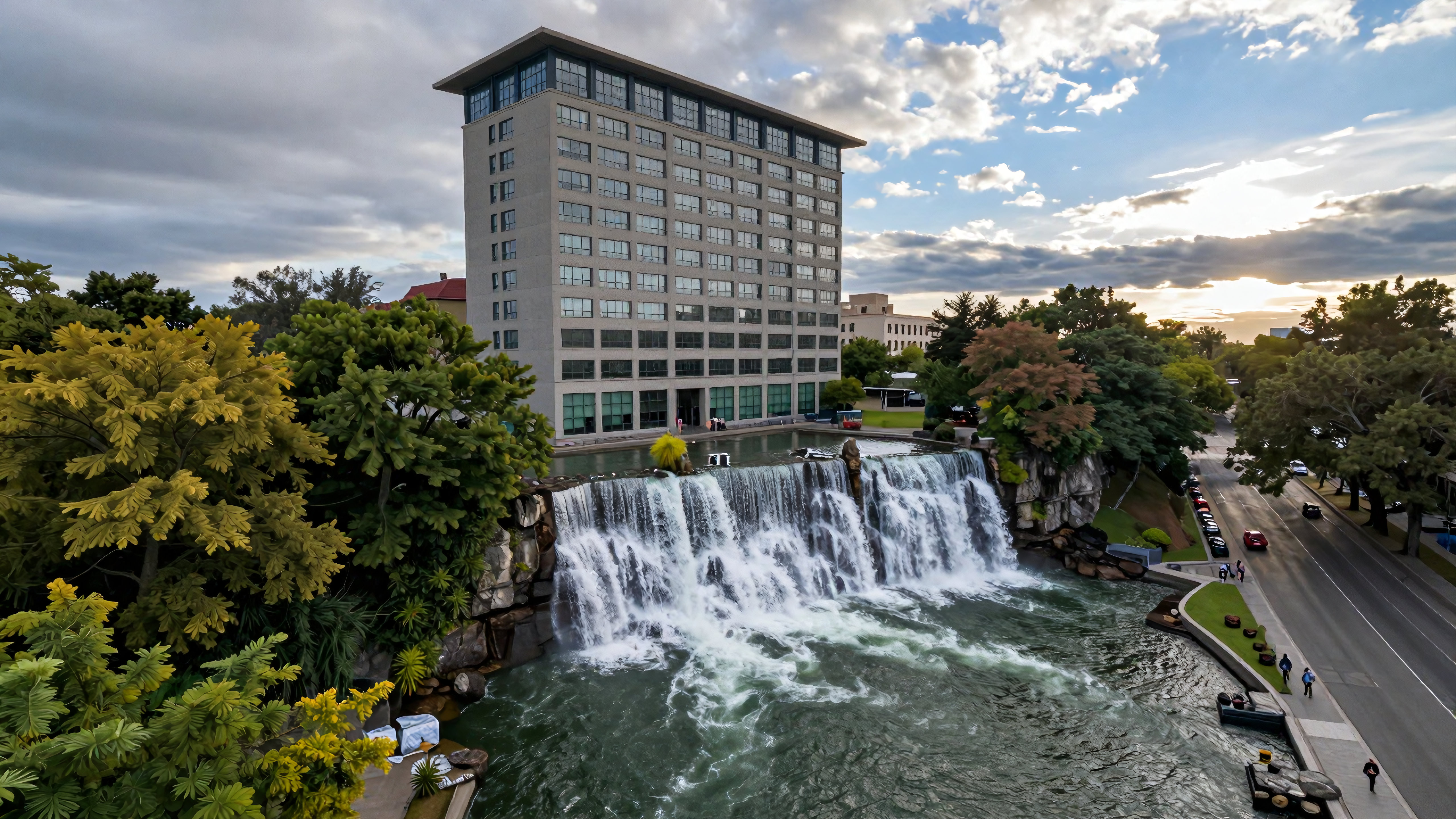Modern building beside scenic waterfall with walking paths and parked cars during sunset