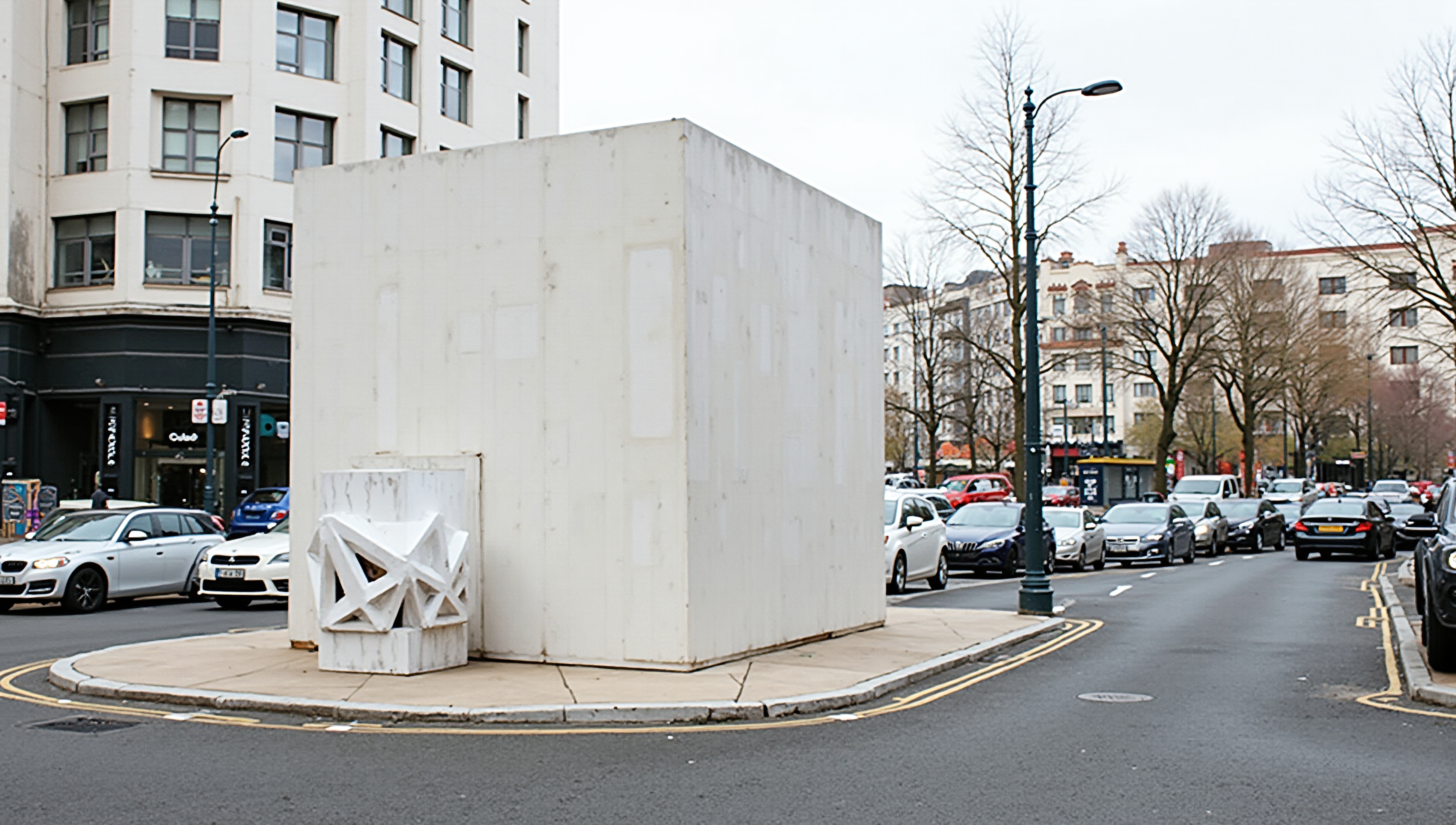 White cubic sculpture standing at a busy urban intersection.