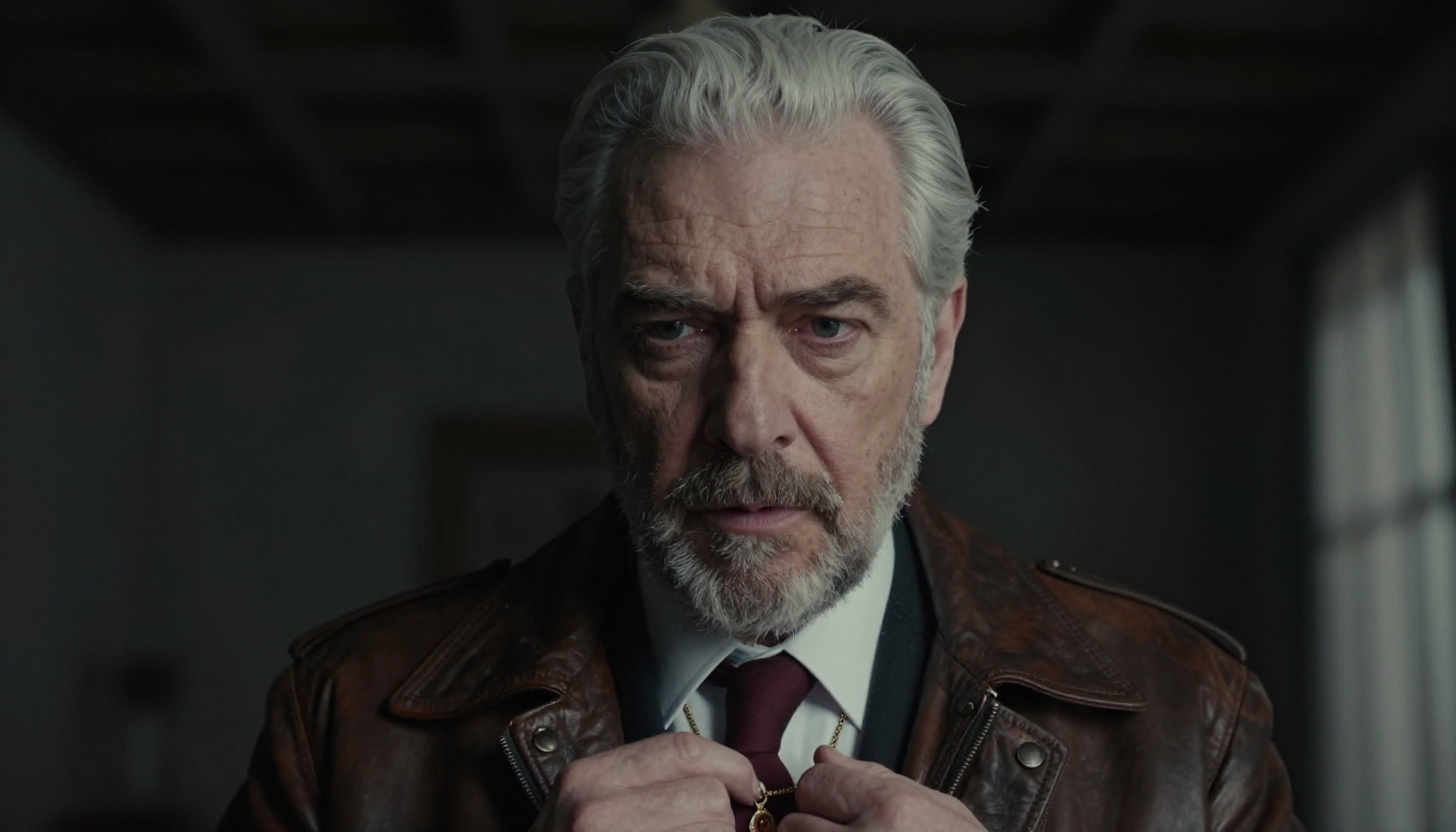 A mature man adjusts his necklace while wearing a leather jacket, conveying depth and contemplation indoors.