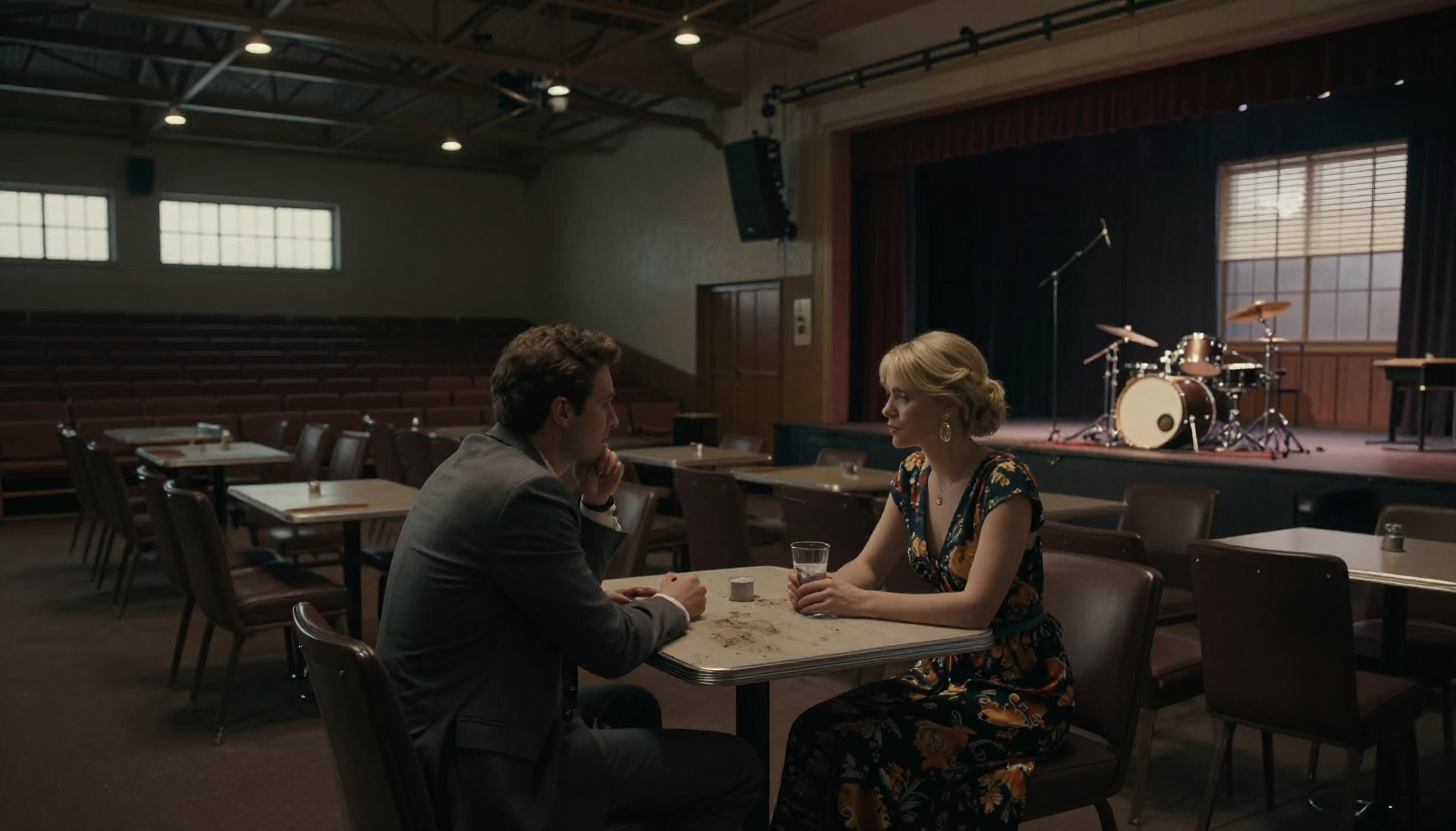 Two people converse in a quiet, dimly lit room with a stage and drum set nearby.