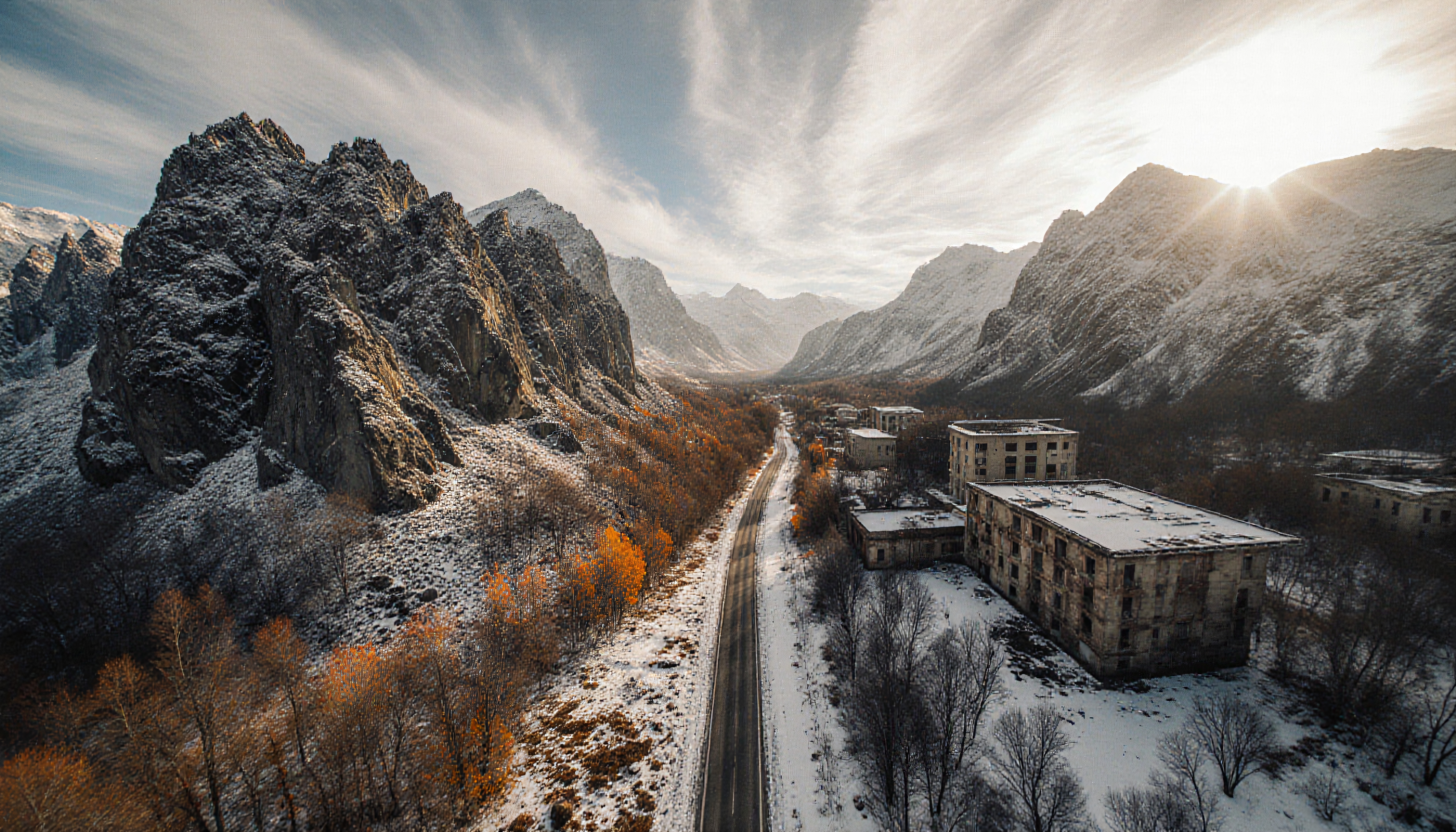 A snow-covered mountain valley with a road flanked by abandoned buildings, brightened by sunlight.