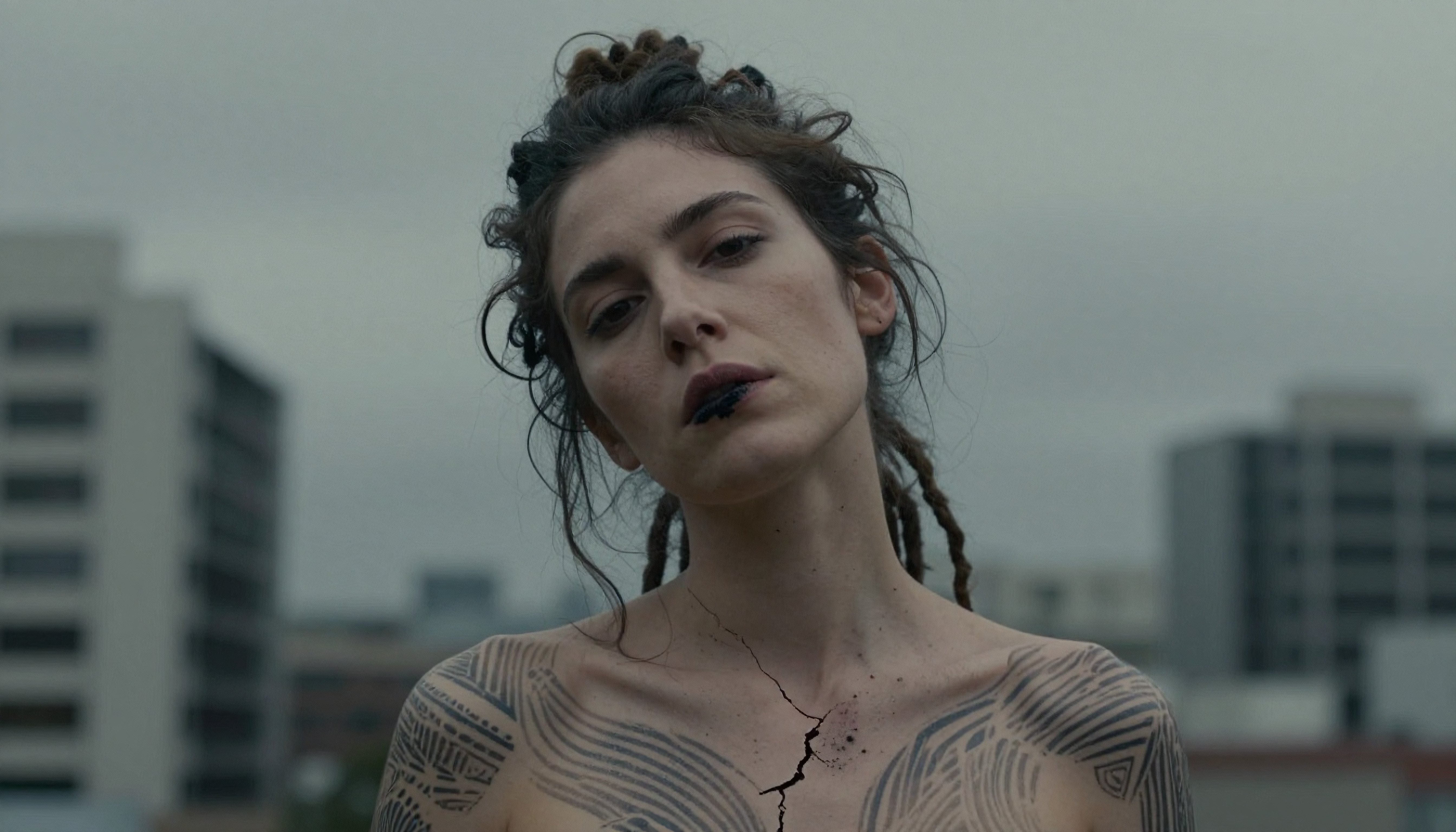 A woman with geometric tattoos and cracked skin effect stands outdoors against a blurred cityscape, under an overcast sky.