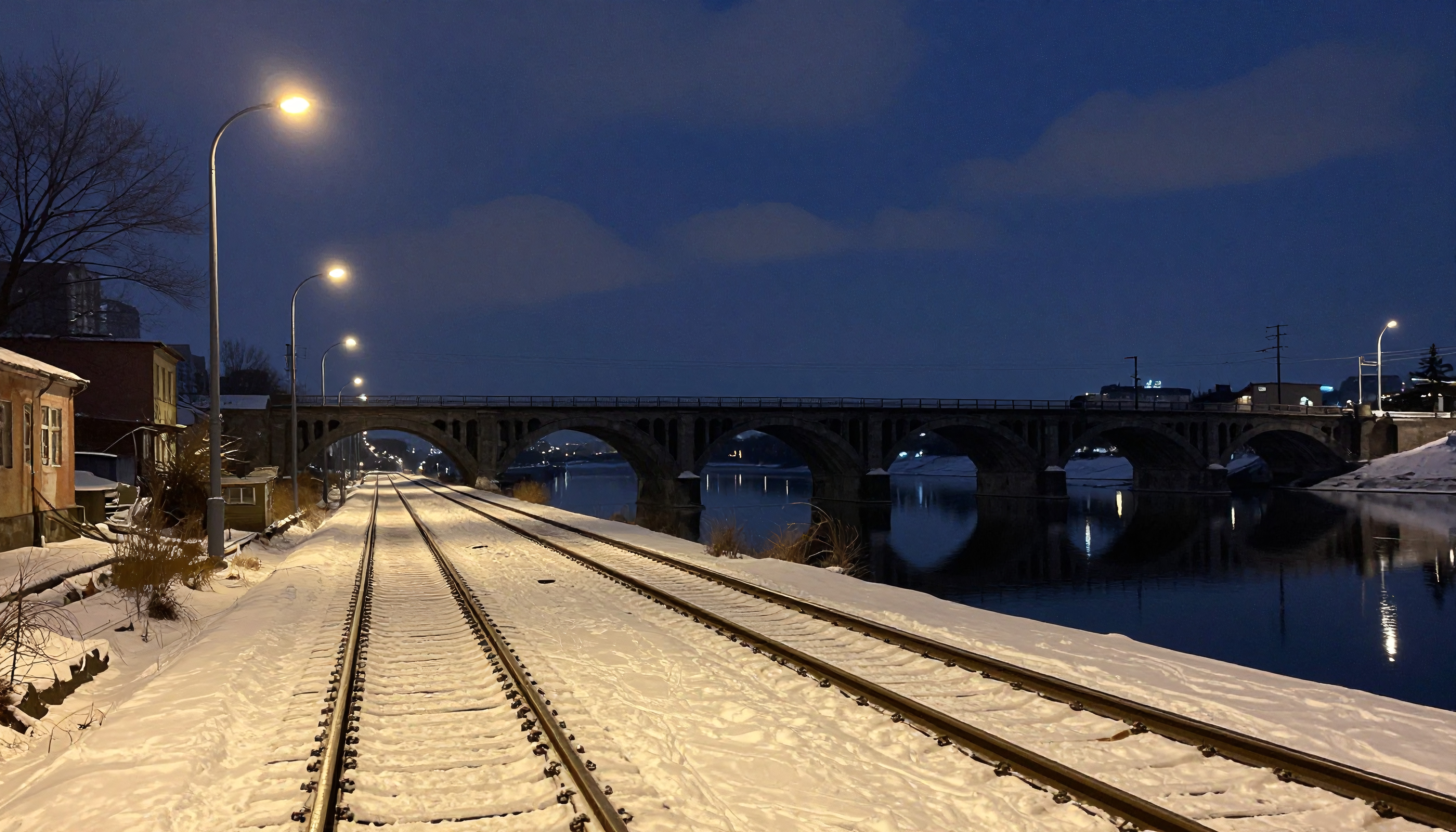 A serene winter evening shows snow-covered railroad tracks leading to a stone bridge, under a dark blue sky, illuminated by streetlights.