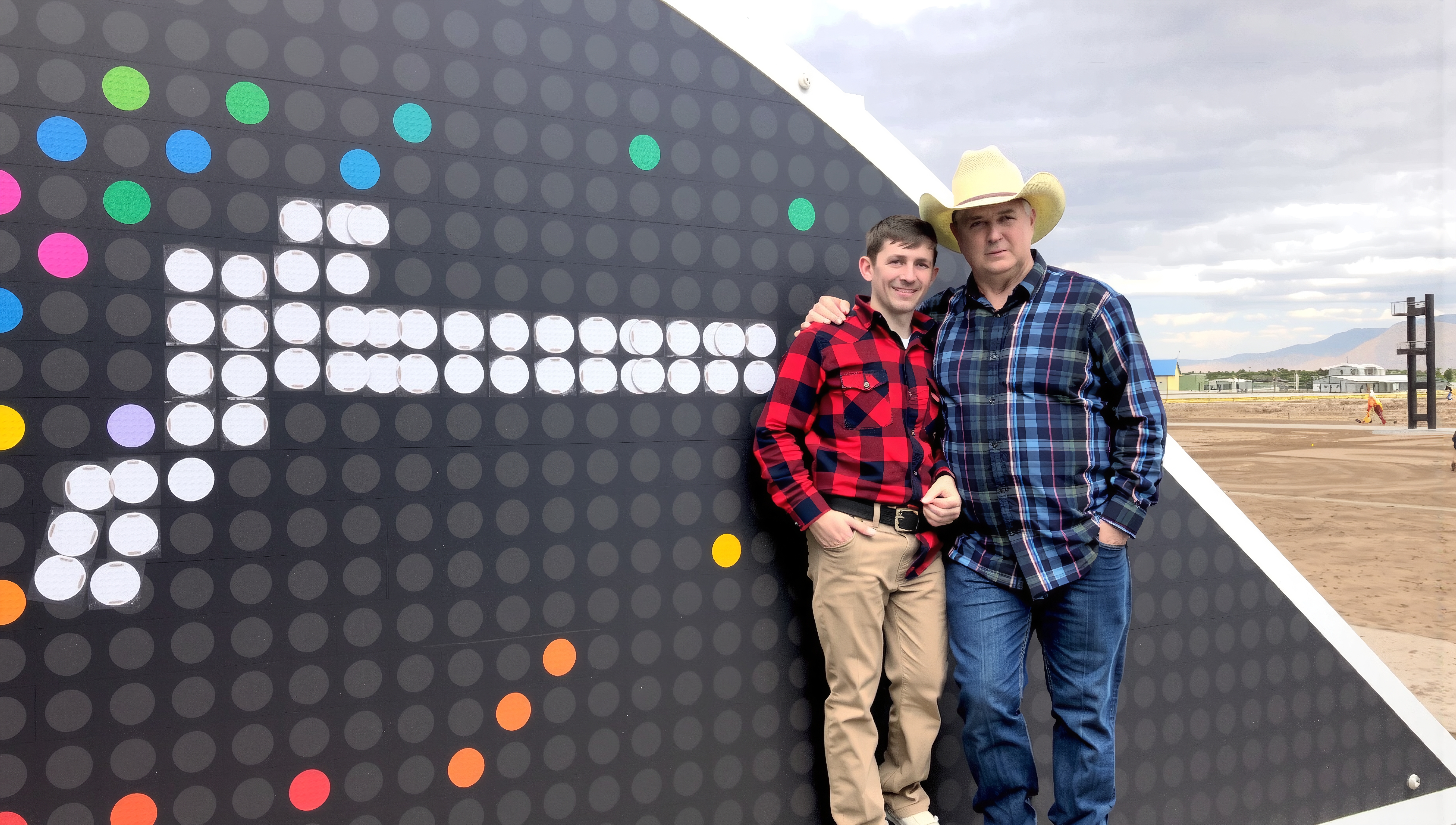 Two men in plaid shirts pose beside a colorful wall outdoors.