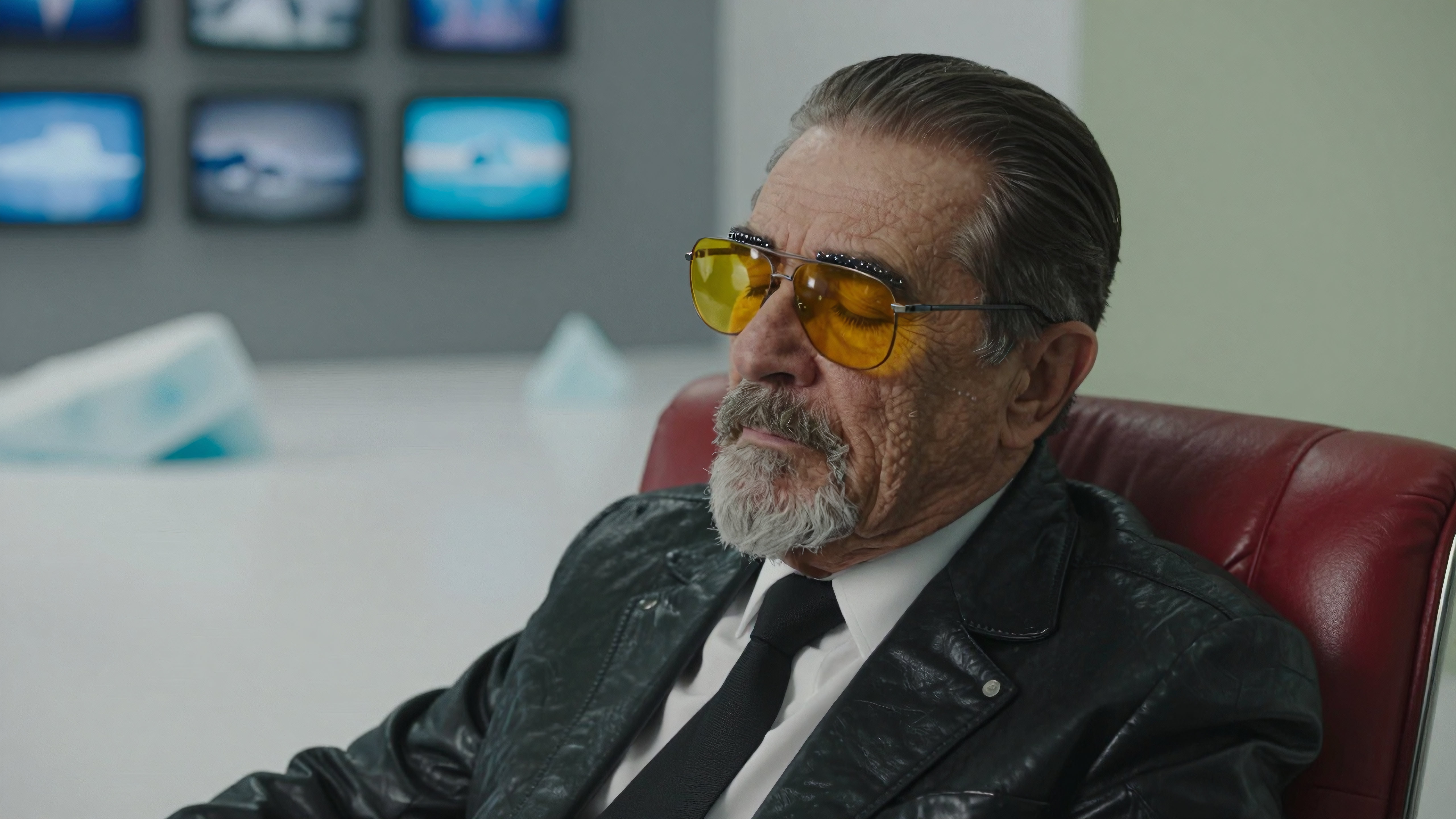 Elderly man wearing sunglasses sitting in a red leather chair surrounded by computer monitors