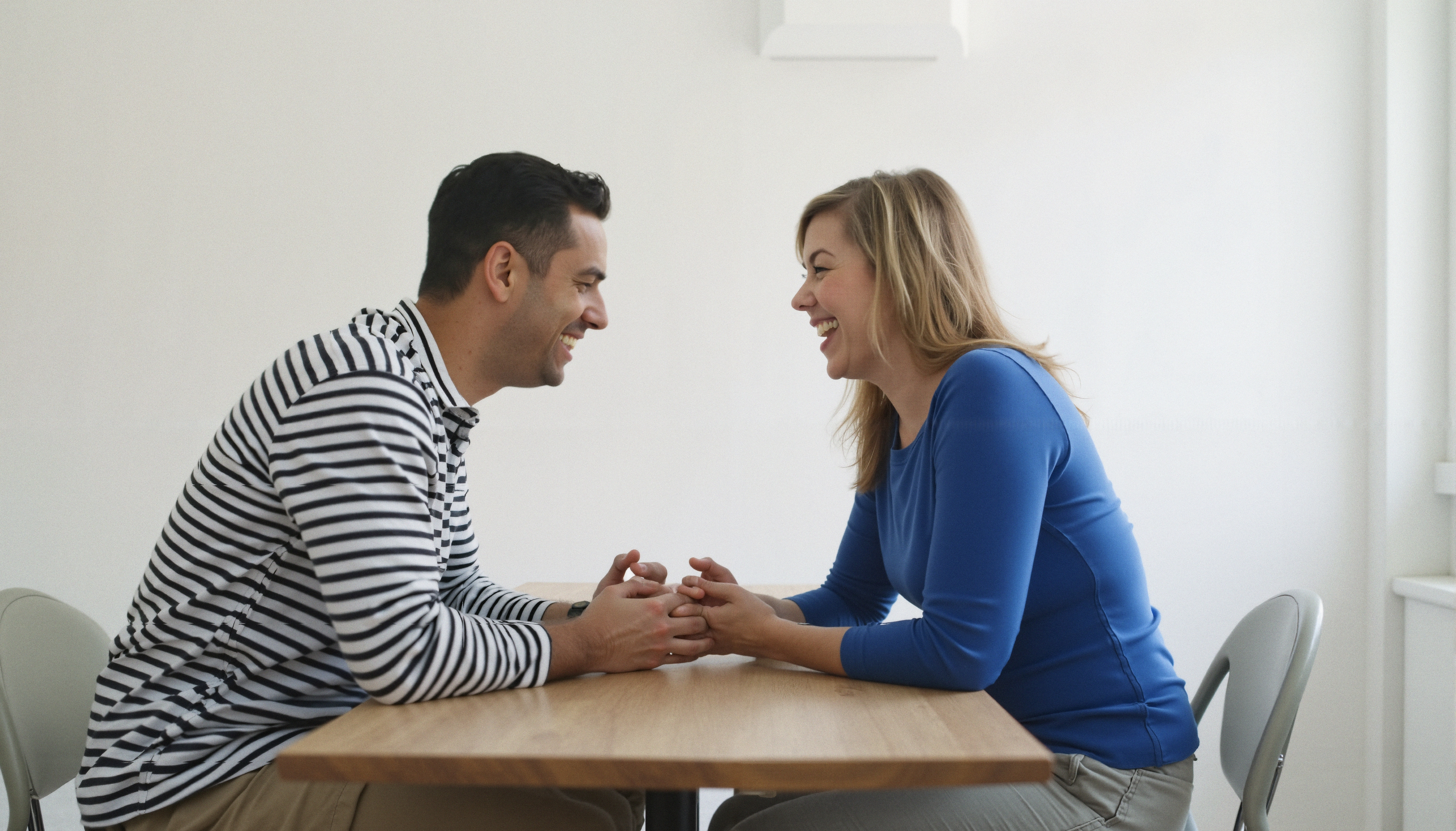 A man and woman happily conversing while holding hands across a table, wearing casual clothing, in a minimalistic setting.