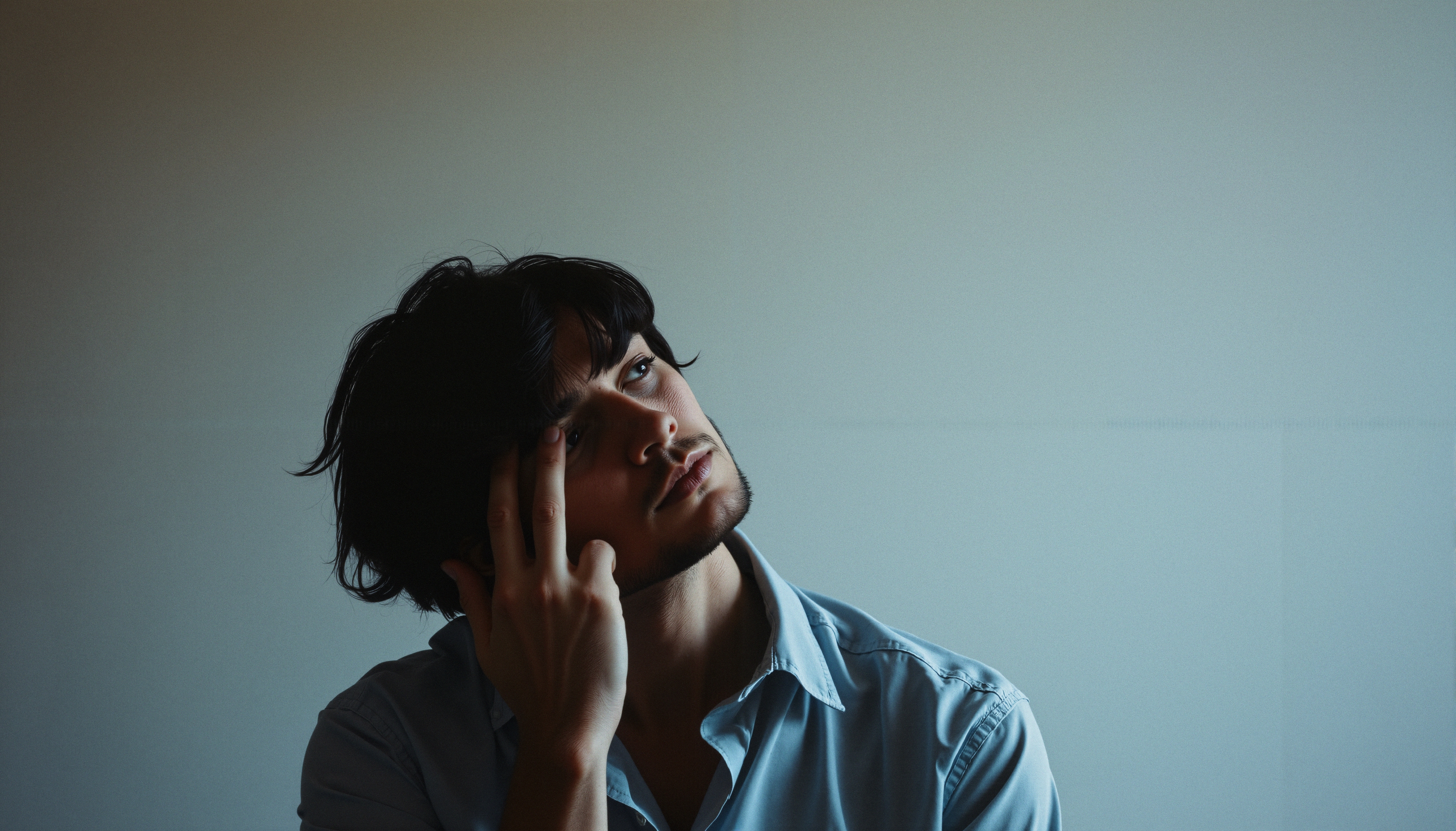 A young person in a light blue shirt gazes thoughtfully, fingers at their temple, immersed in quiet reflection.