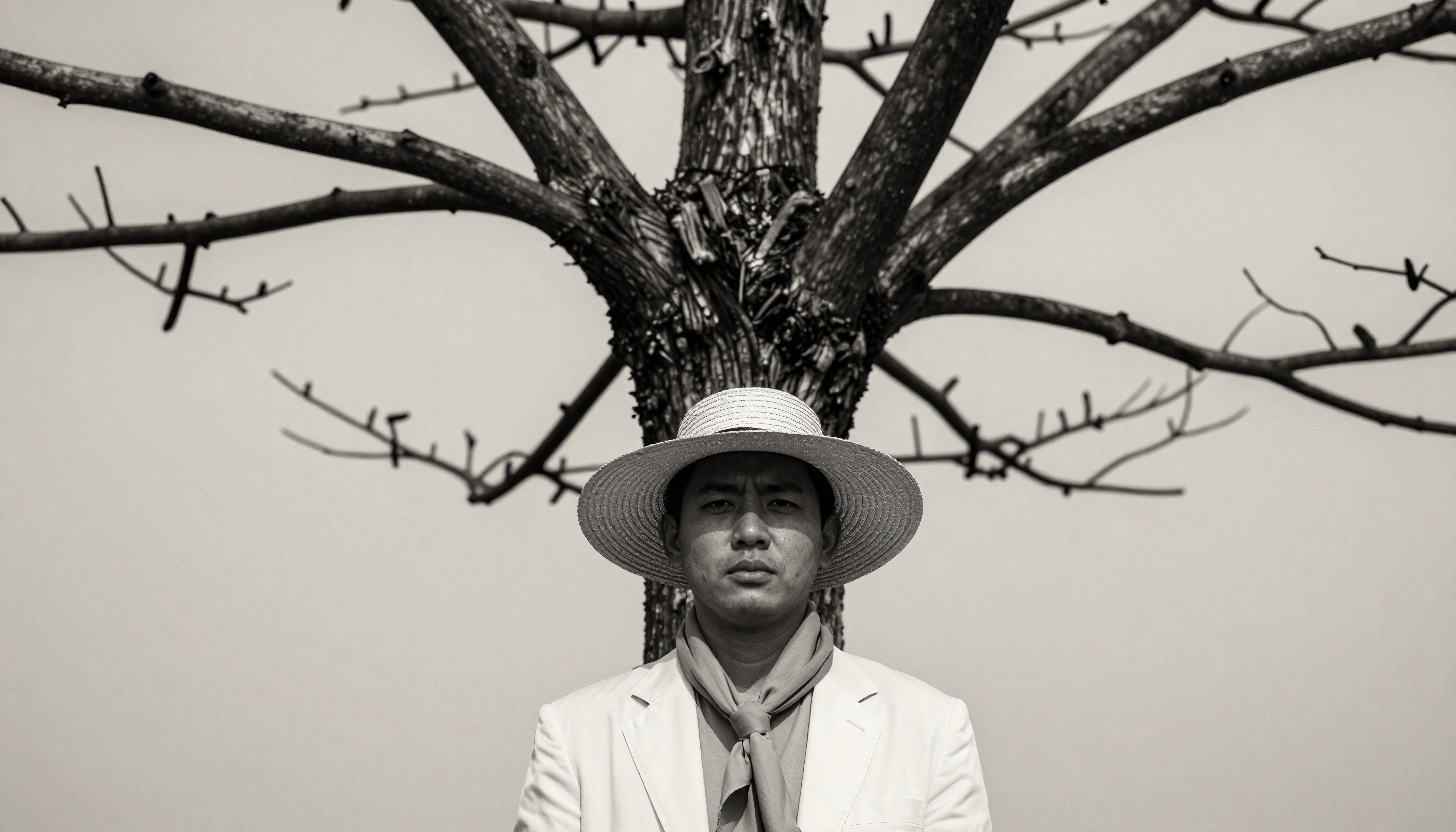 A man in a straw hat stands symmetrically aligned with a tree behind him, creating a striking composition in black and white.