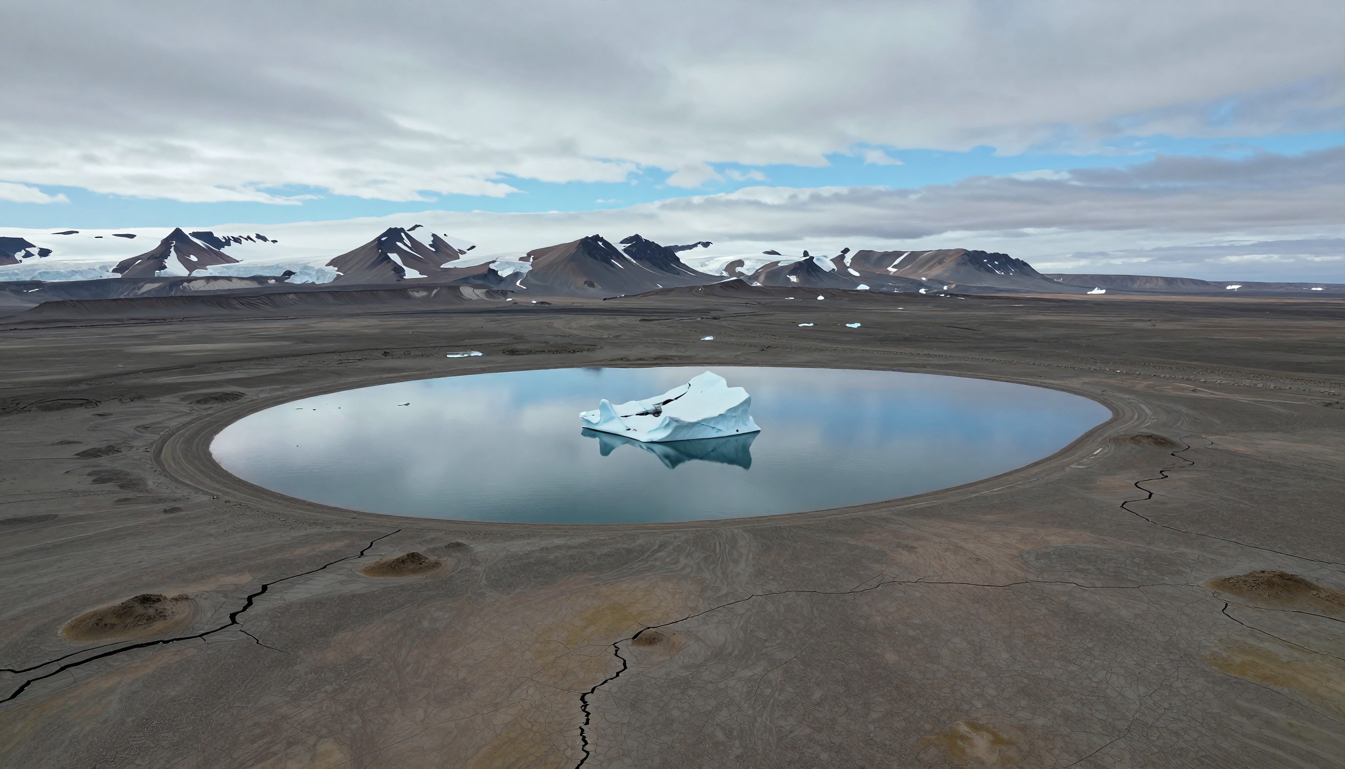 An iceberg floats in a circular lake surrounded by barren land and snow-capped mountains.