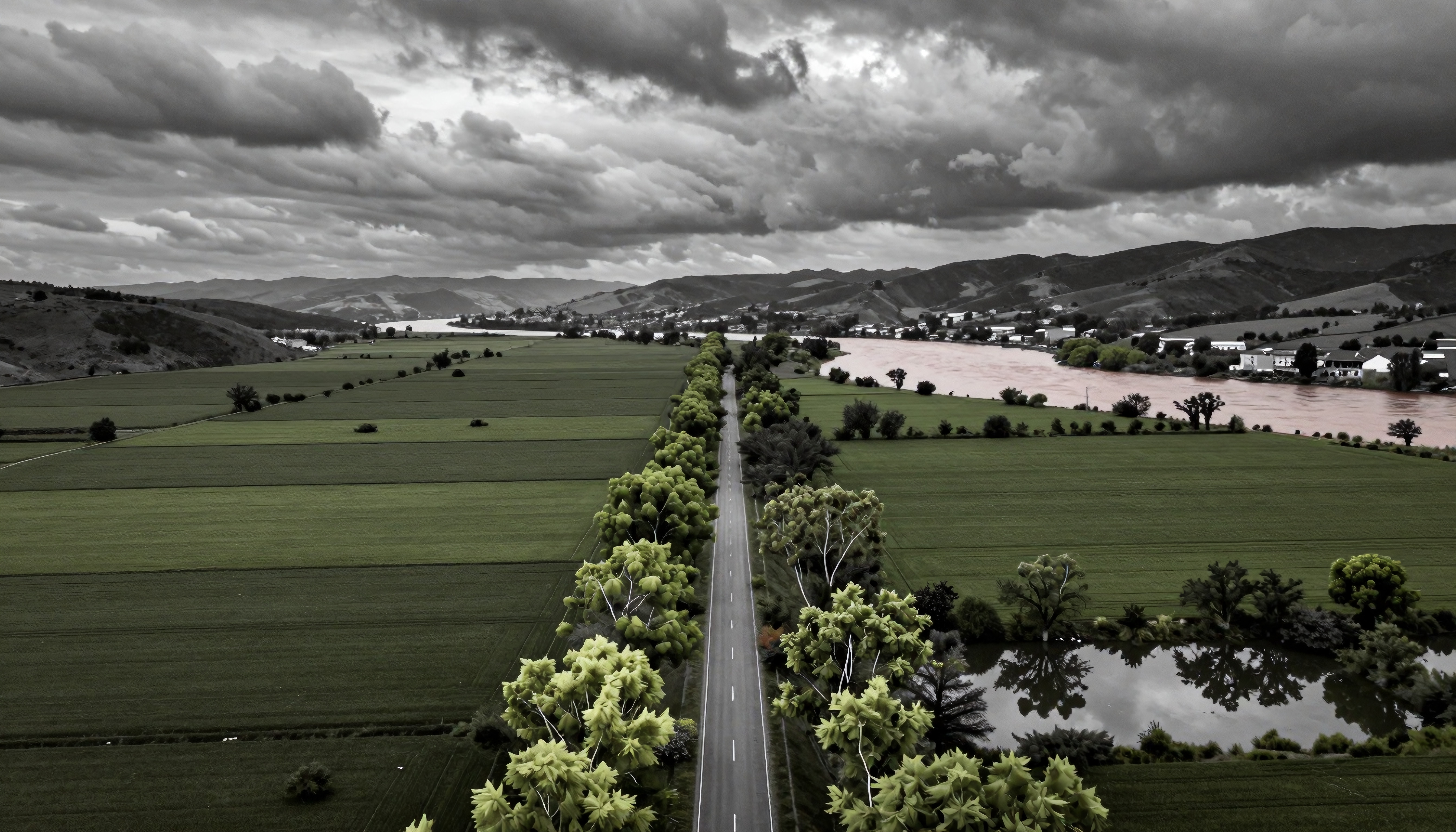An aerial view of a road cutting through green fields, lined with vibrant trees, under a sky of dark storm clouds and beside a winding river.