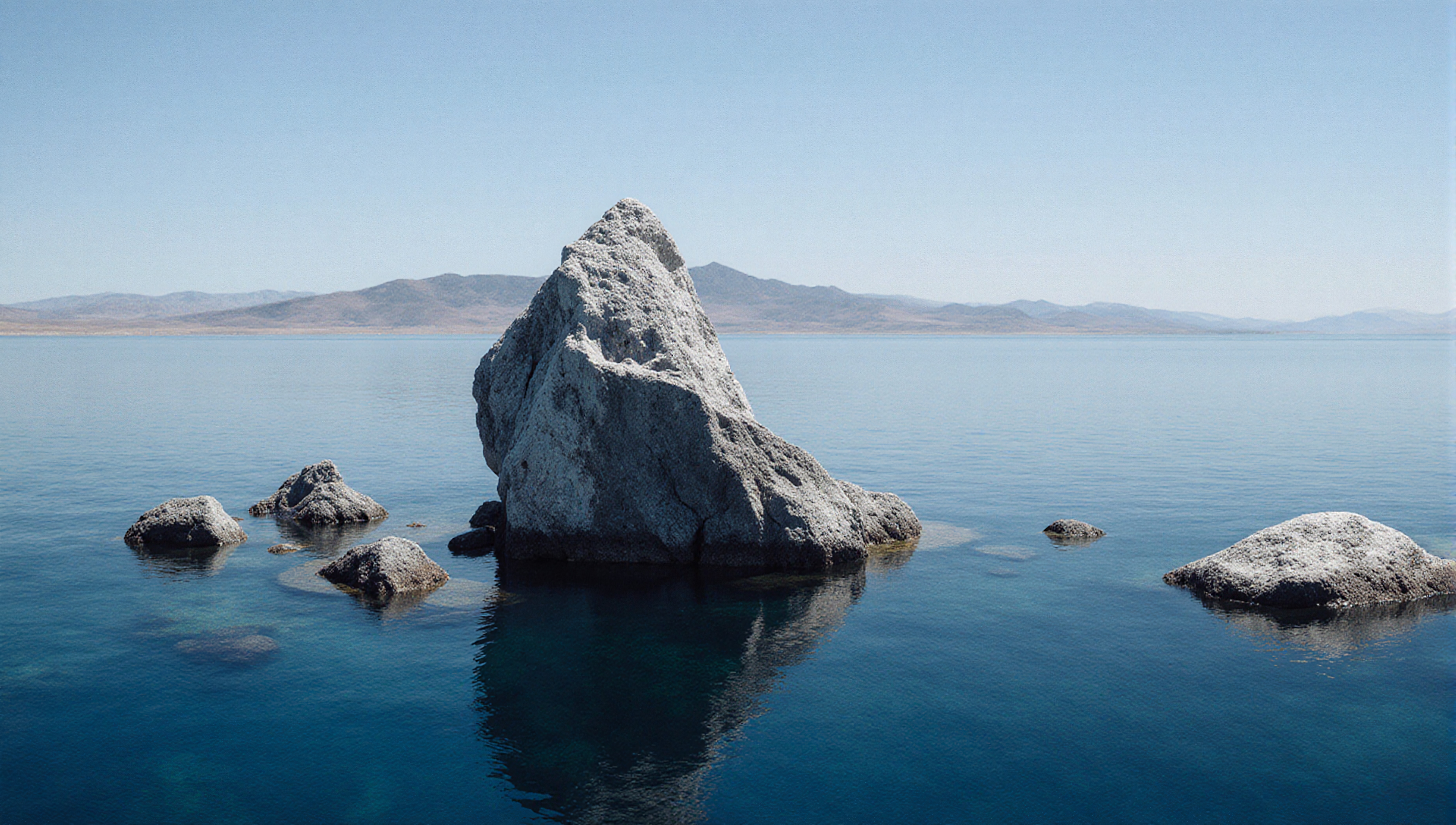 Large rock and smaller rocks partly submerged in a calm, blue body of water with distant hills under a clear sky.