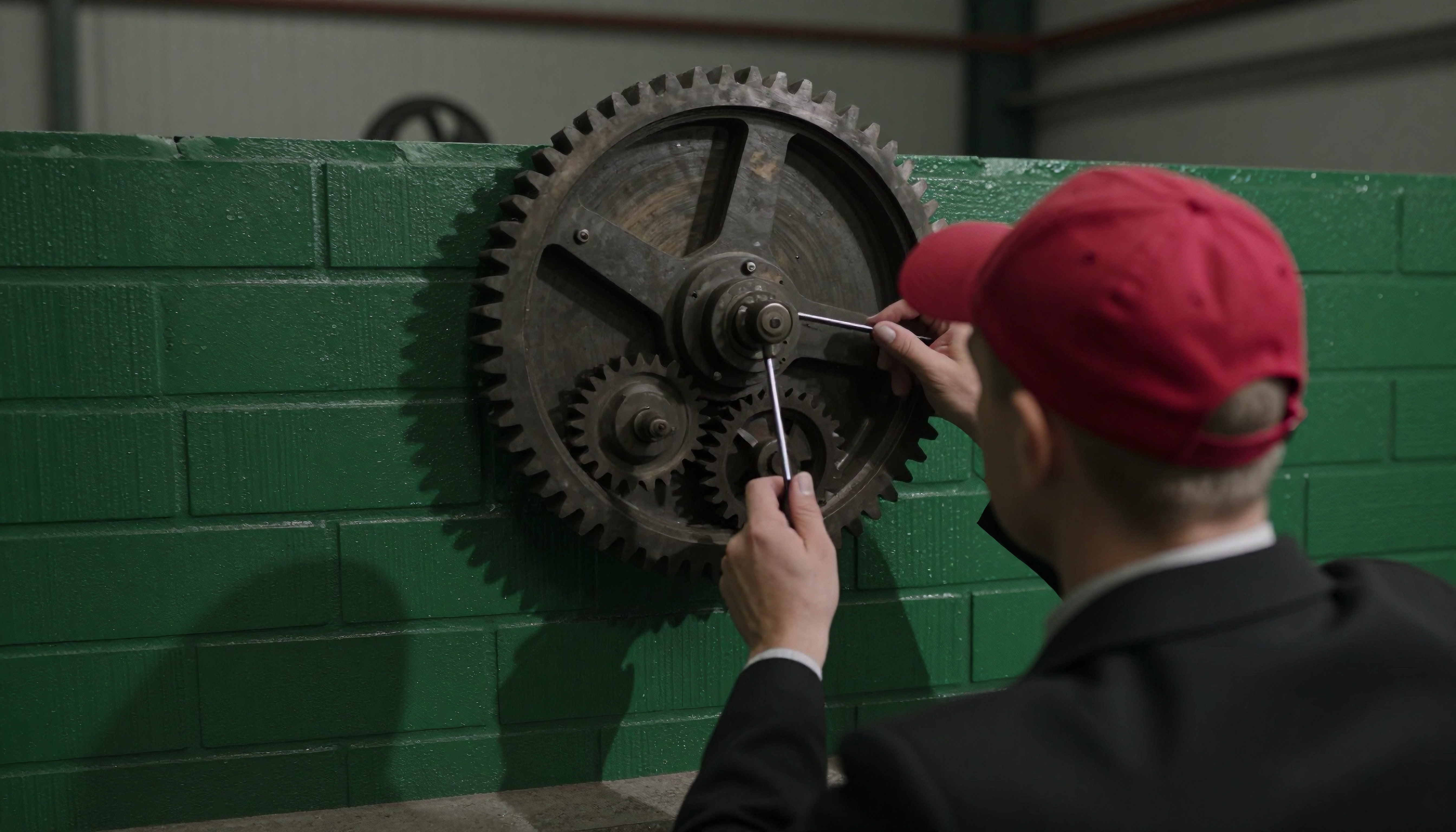 A man adjusts mechanical gears on a green brick wall.