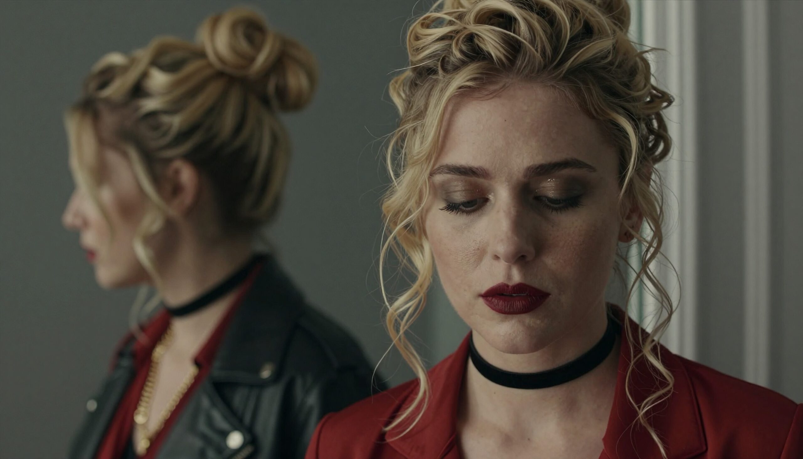 A woman in red contemplates by a mirror wearing a choker and leather.