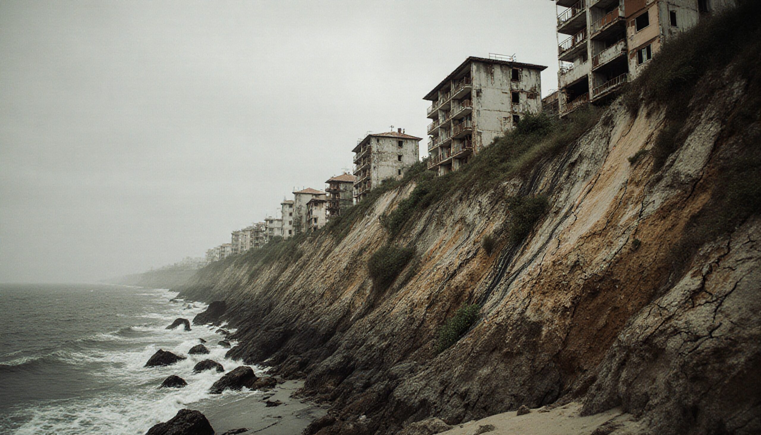 Abandoned buildings perched on eroded cliffside with waves crashing below under an overcast sky.