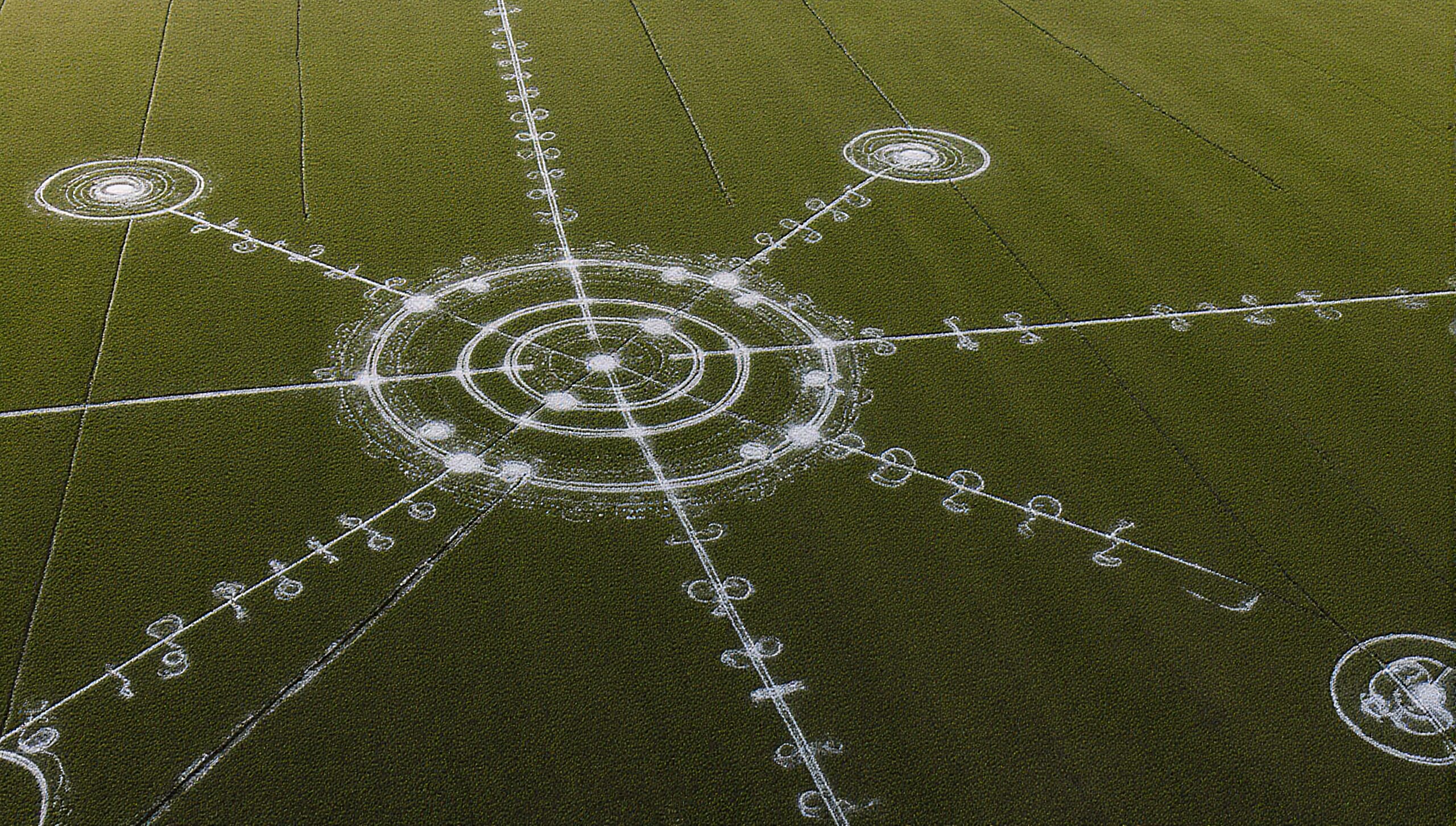 Aerial view of an intricate crop circle design in a green field, featuring concentric geometric patterns and connecting lines.