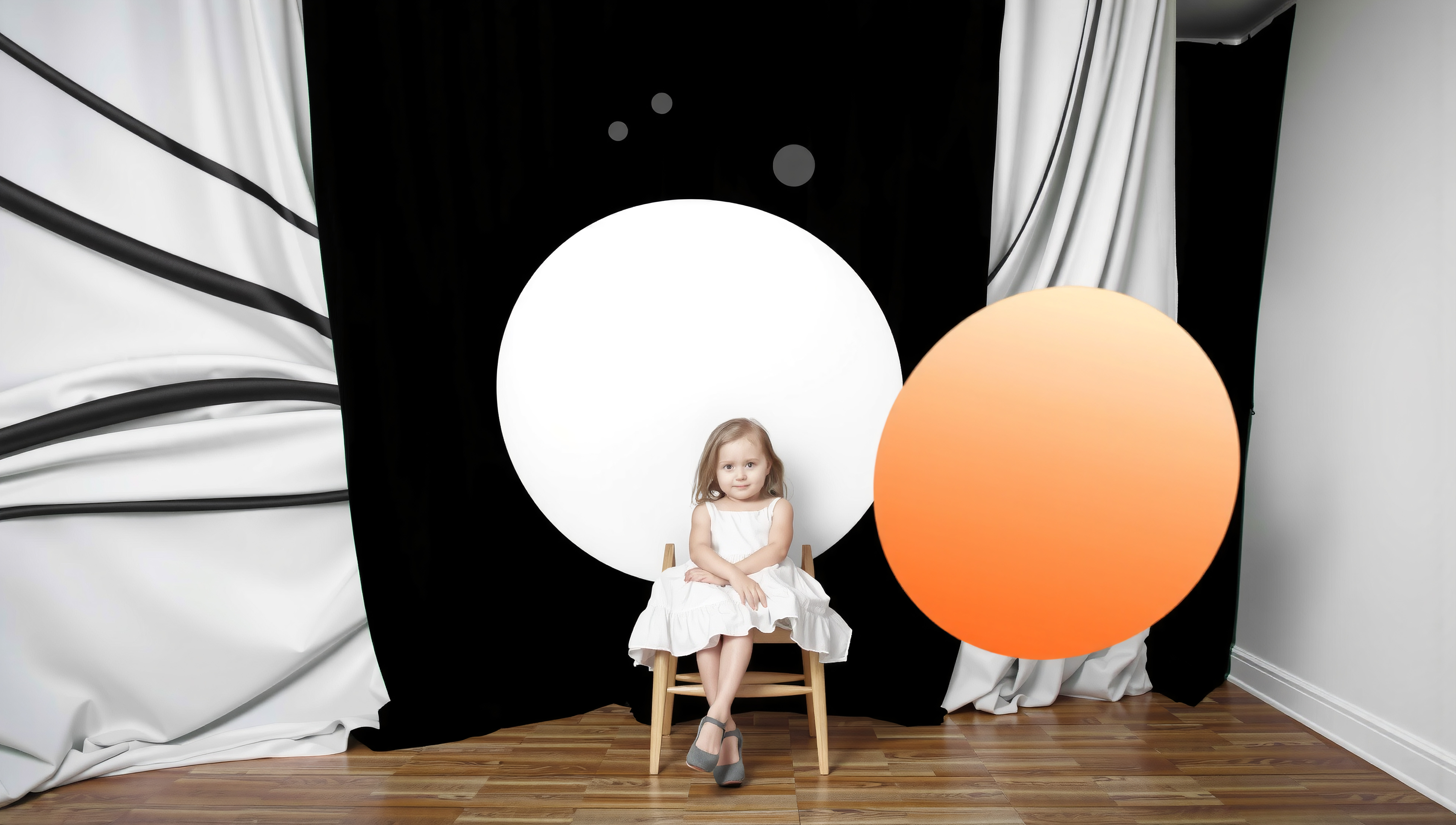A young girl in a white dress sits calmly on a wooden chair against a backdrop of black and white circles with an overlapping orange element, on a polished wooden floor.